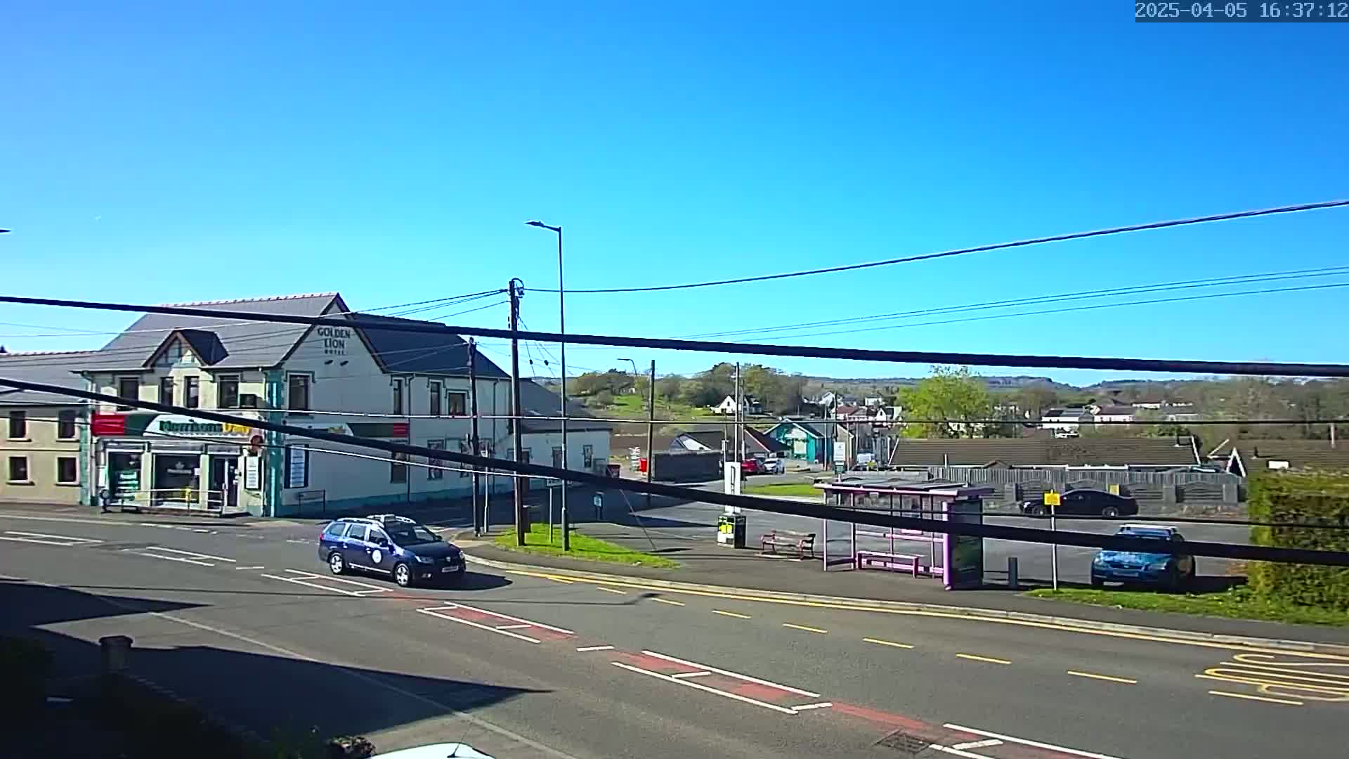 Under a clear blue sky, a street scene shows a two-story building with a smaller shop attached, a bus stop, several cars, and a background of low hills and greenery.