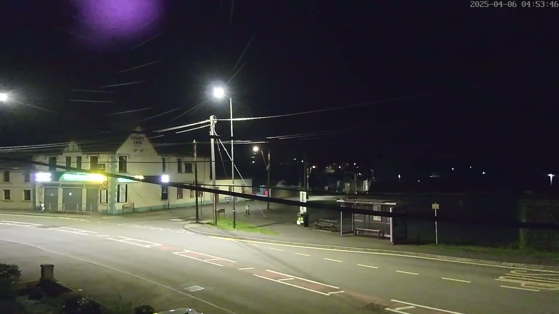A nighttime street scene shows a two-story building, a bus stop, and a road intersection illuminated by streetlights under a dark sky.