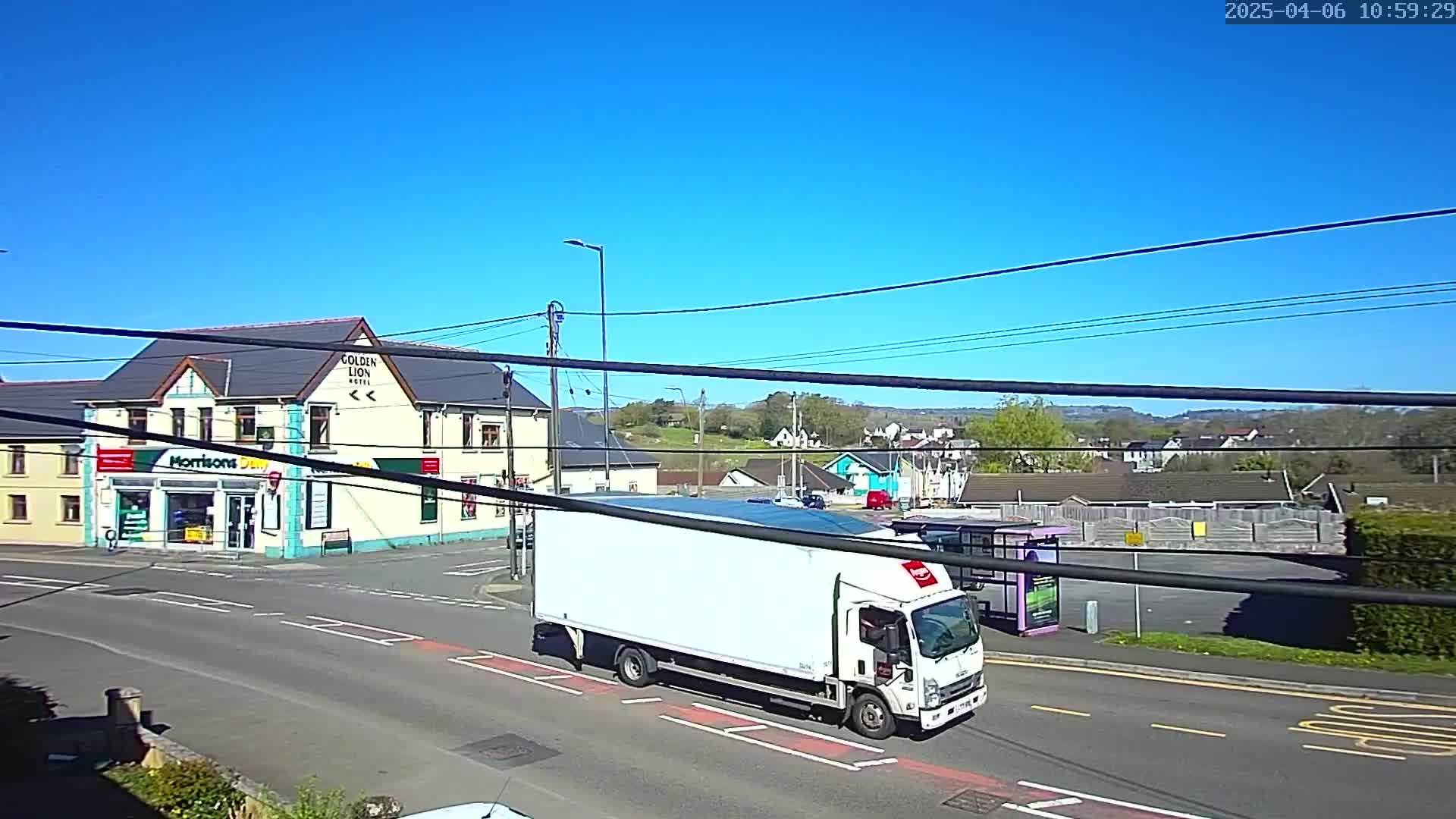 A white delivery truck drives past a corner building with a shop and hotel, on a sunny day.