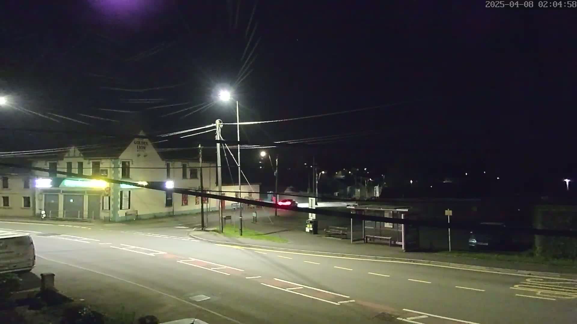A nighttime street scene shows a road intersection with a building, a bus shelter, and several cars under a dark, clear sky.