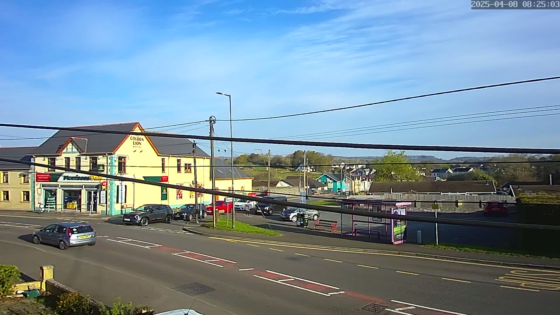 A sunny day reveals a street scene with a yellow building, several cars, and a bus shelter, all under a mostly clear sky.