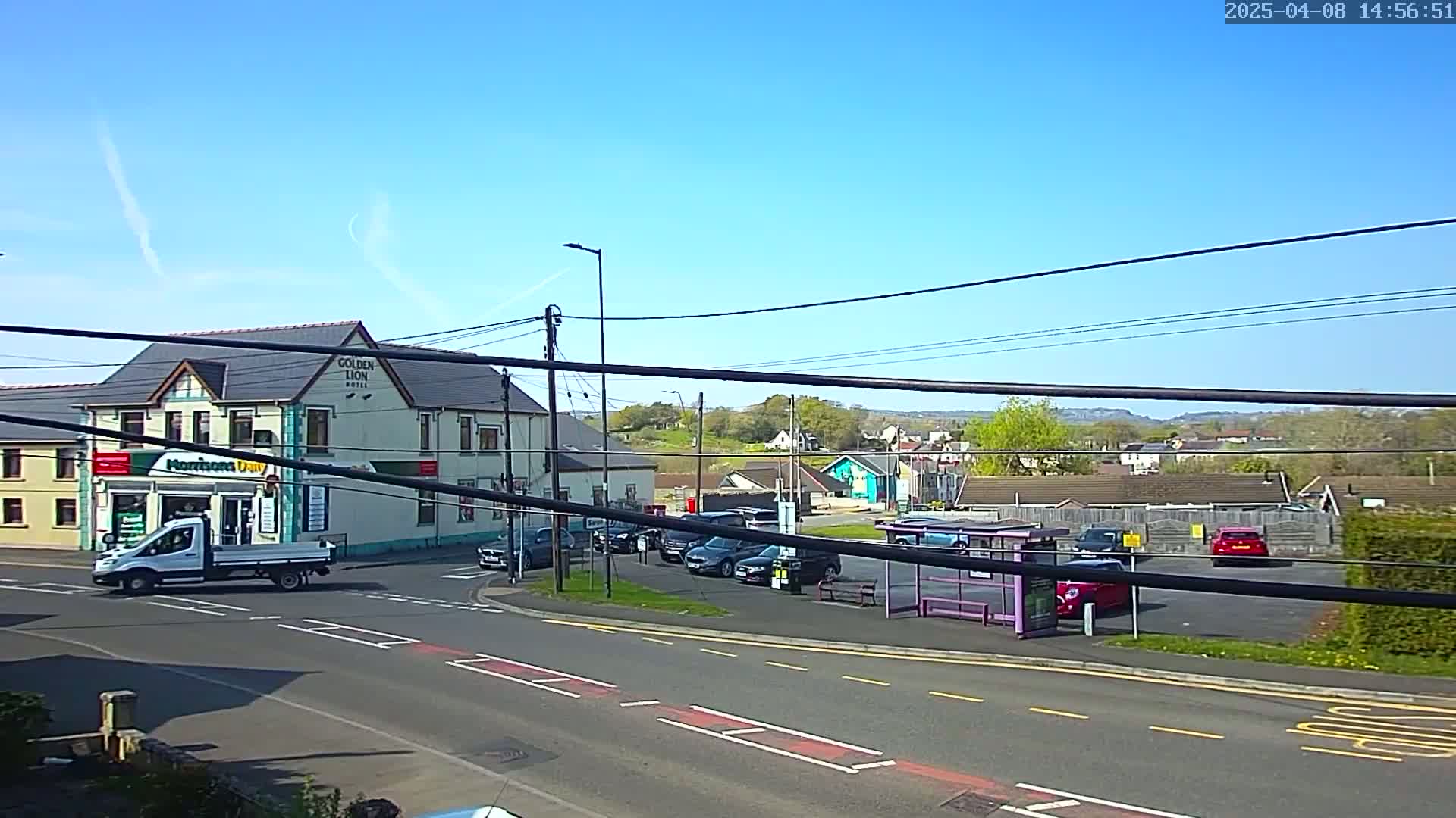 A sunny day reveals a street scene with a two-story building, several parked cars, a bus stop, and a small white delivery truck at an intersection.
