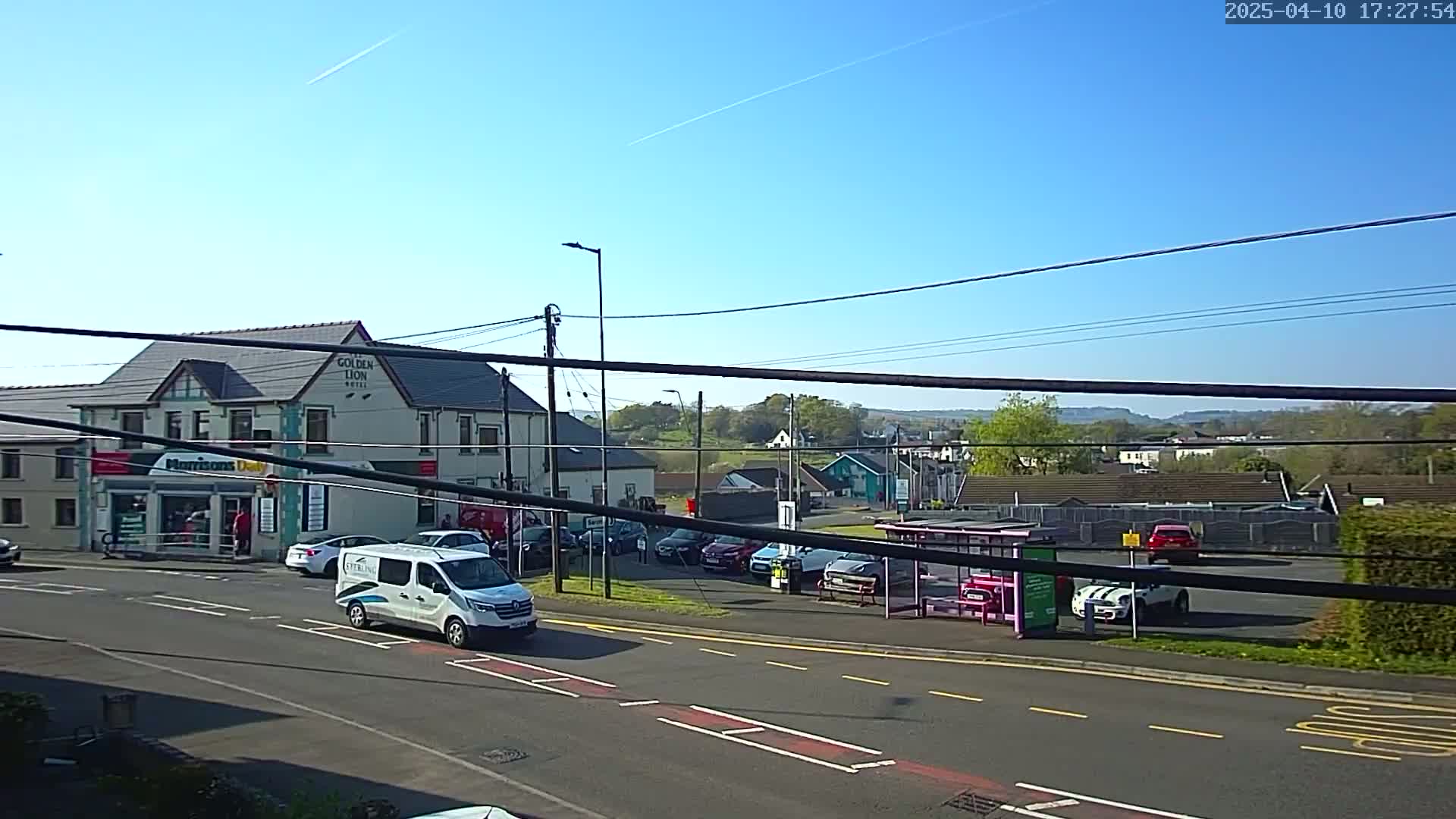 A sunny day reveals a street scene with a hotel, shops, parked cars, and a bus shelter, all set against a backdrop of rolling hills.