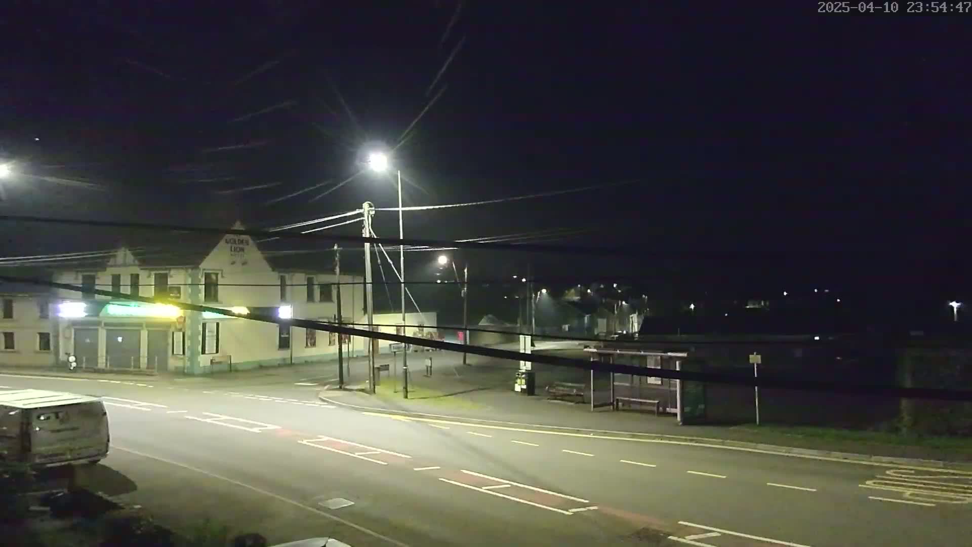 A nighttime street scene shows a mostly empty road, a building, a bus stop, and several streetlights under a clear, dark sky.