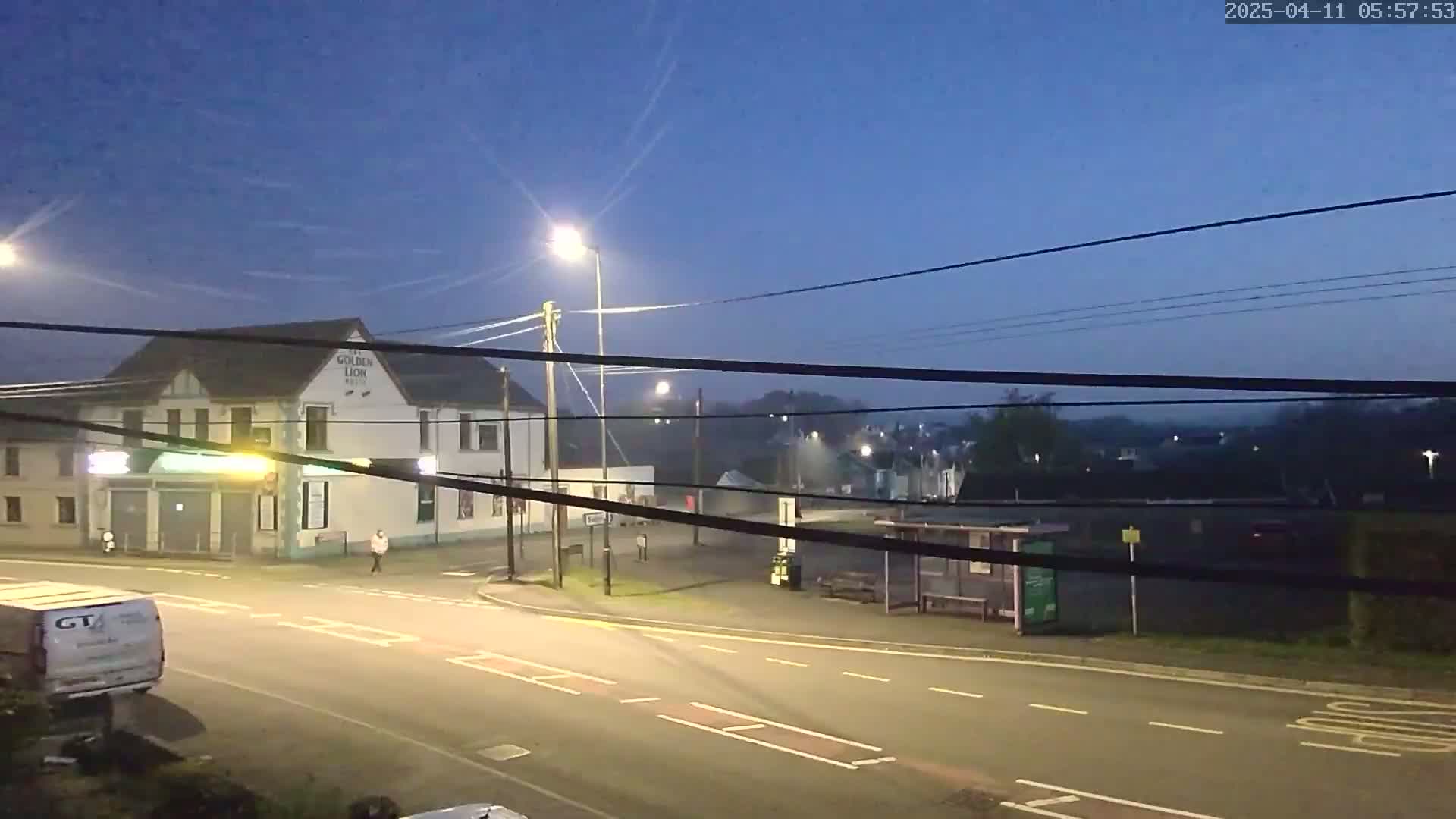 A dimly lit street scene at twilight shows a pub, a bus shelter, a few pedestrians, and some vehicles under a clear, dark sky.