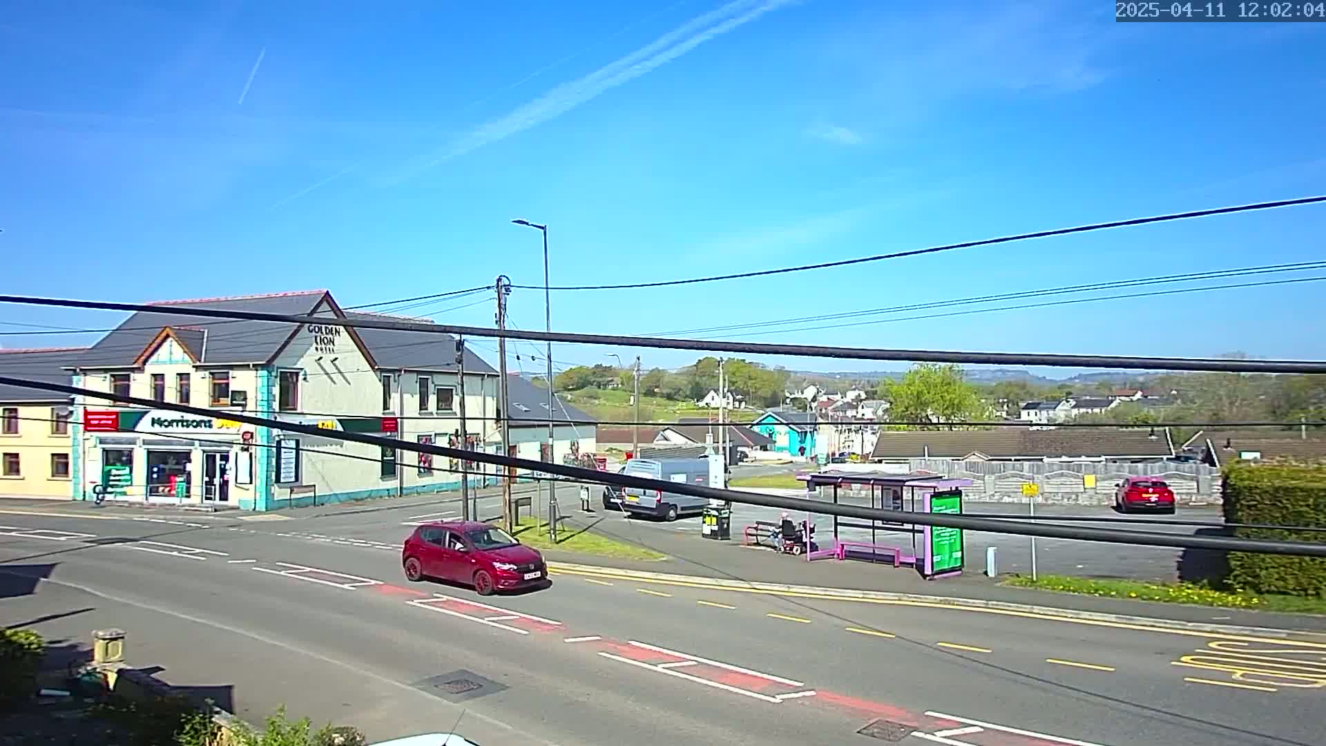 A sunny day reveals a street scene with a red car, a white van, and a bus stop near buildings that include a pub and a shop.