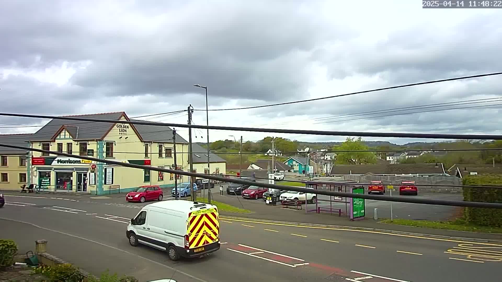 A white van drives past a crossroads in front of a hotel and shops under an overcast sky.