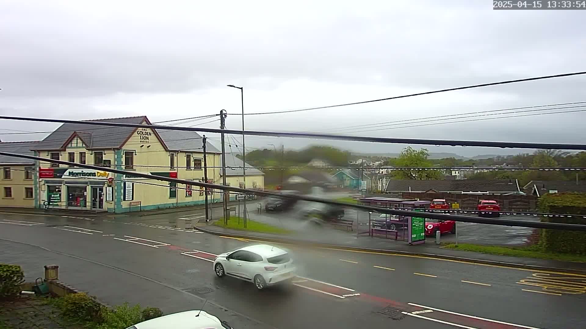 A rainy day scene shows a street intersection with a yellow building housing a shop and pub, several cars, and a bus stop.