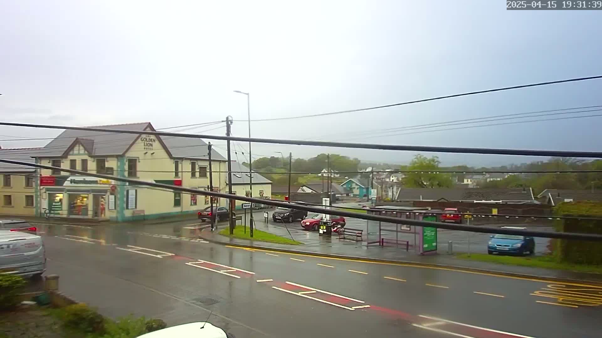 A wet, overcast street scene shows a corner building with a bus stop and several cars parked nearby.