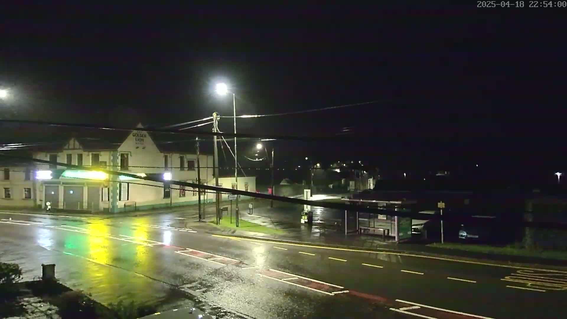 A wet, nighttime street scene shows a building with lights on, a bus stop, and streetlights illuminating a rain-slicked road.