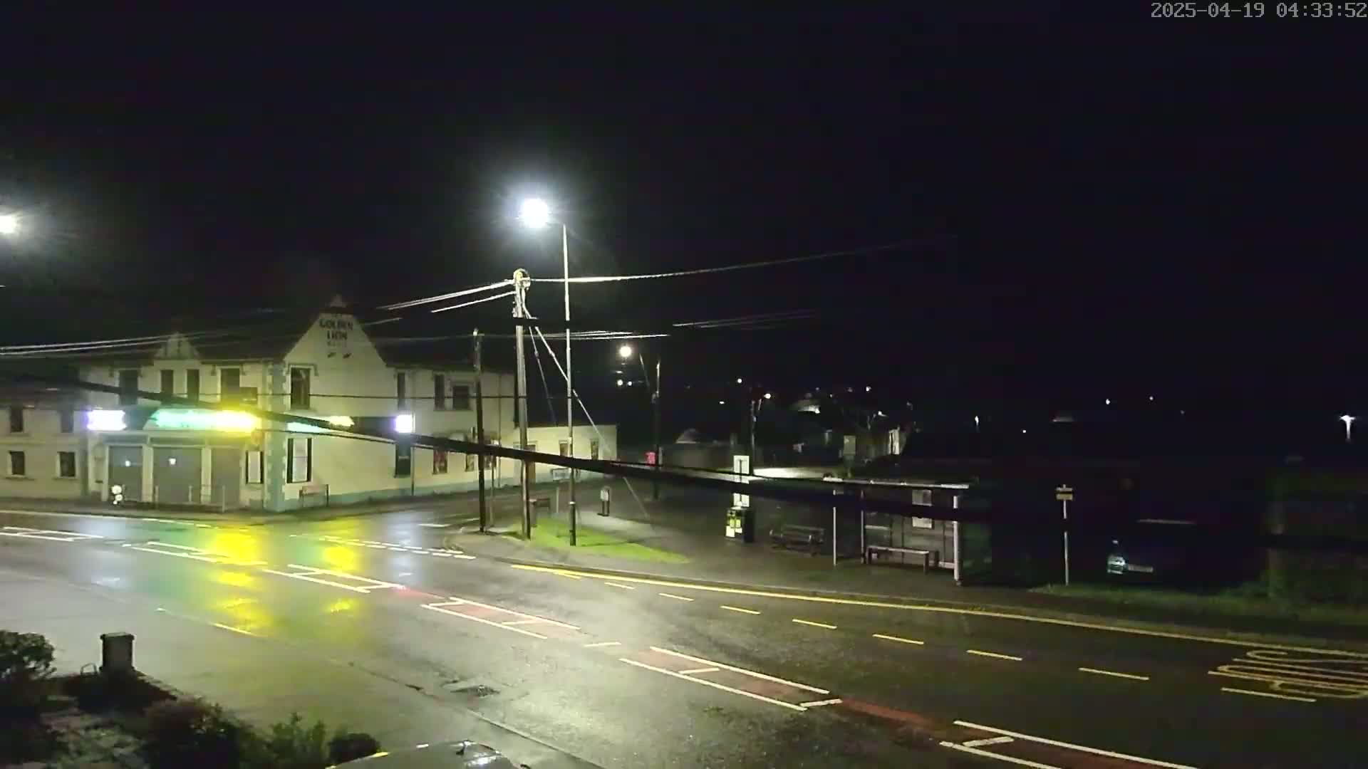 A wet, nighttime street scene shows a building with lights on, a bus stop, and streetlights illuminating a mostly empty road.