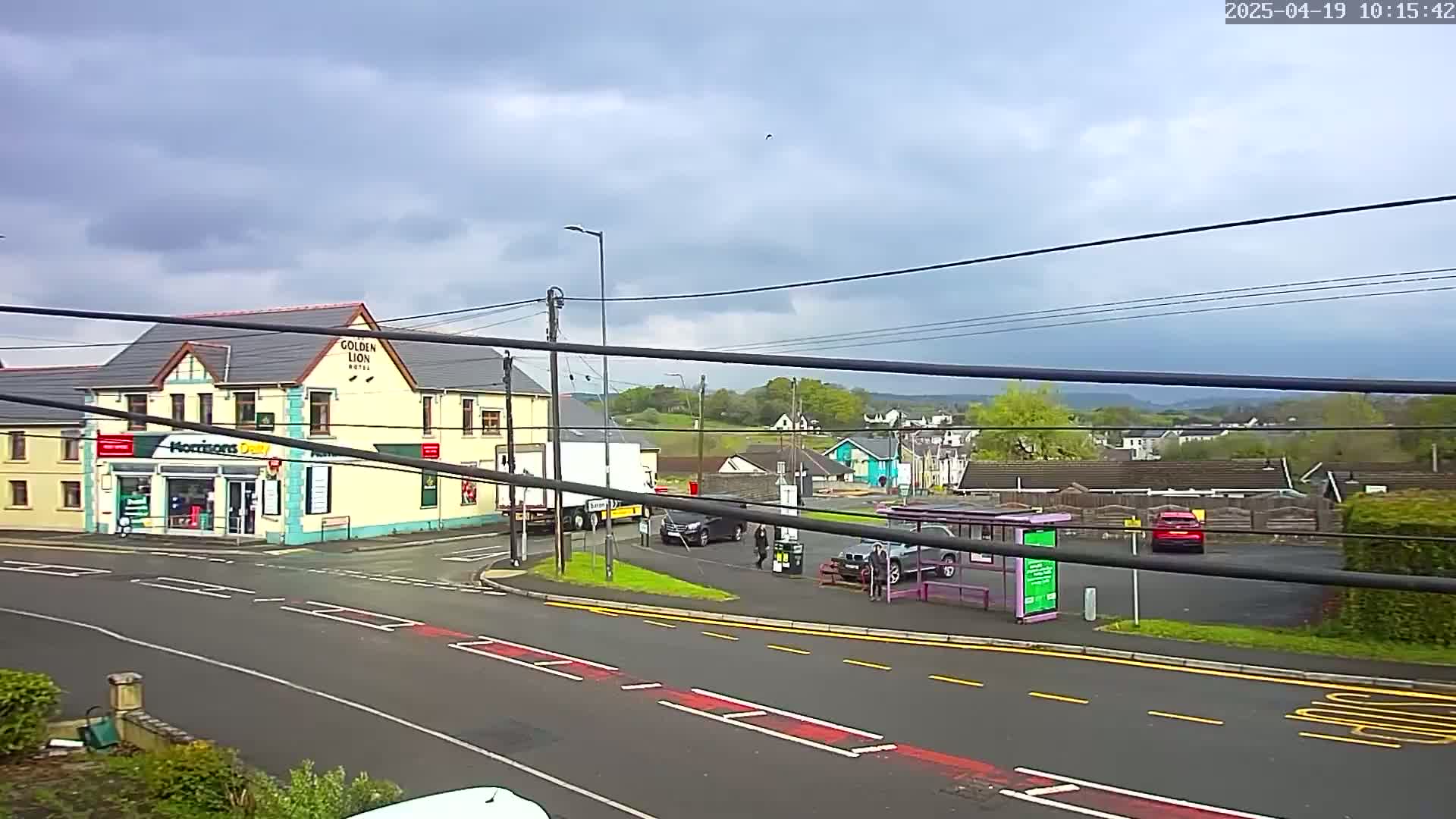 A mostly cloudy day reveals a street scene with a yellow building, a bus stop, several cars, and a large truck, all situated near a road intersection.