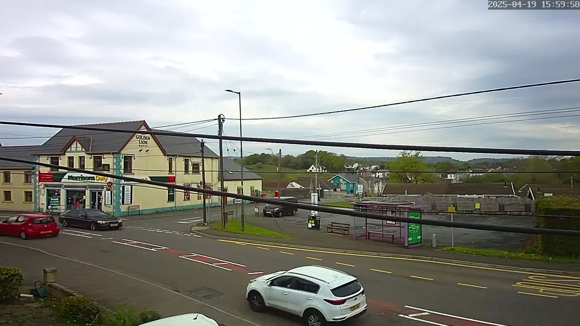 A street scene on an overcast day shows a yellow building, a bus stop, several cars, and green hills in the distance.