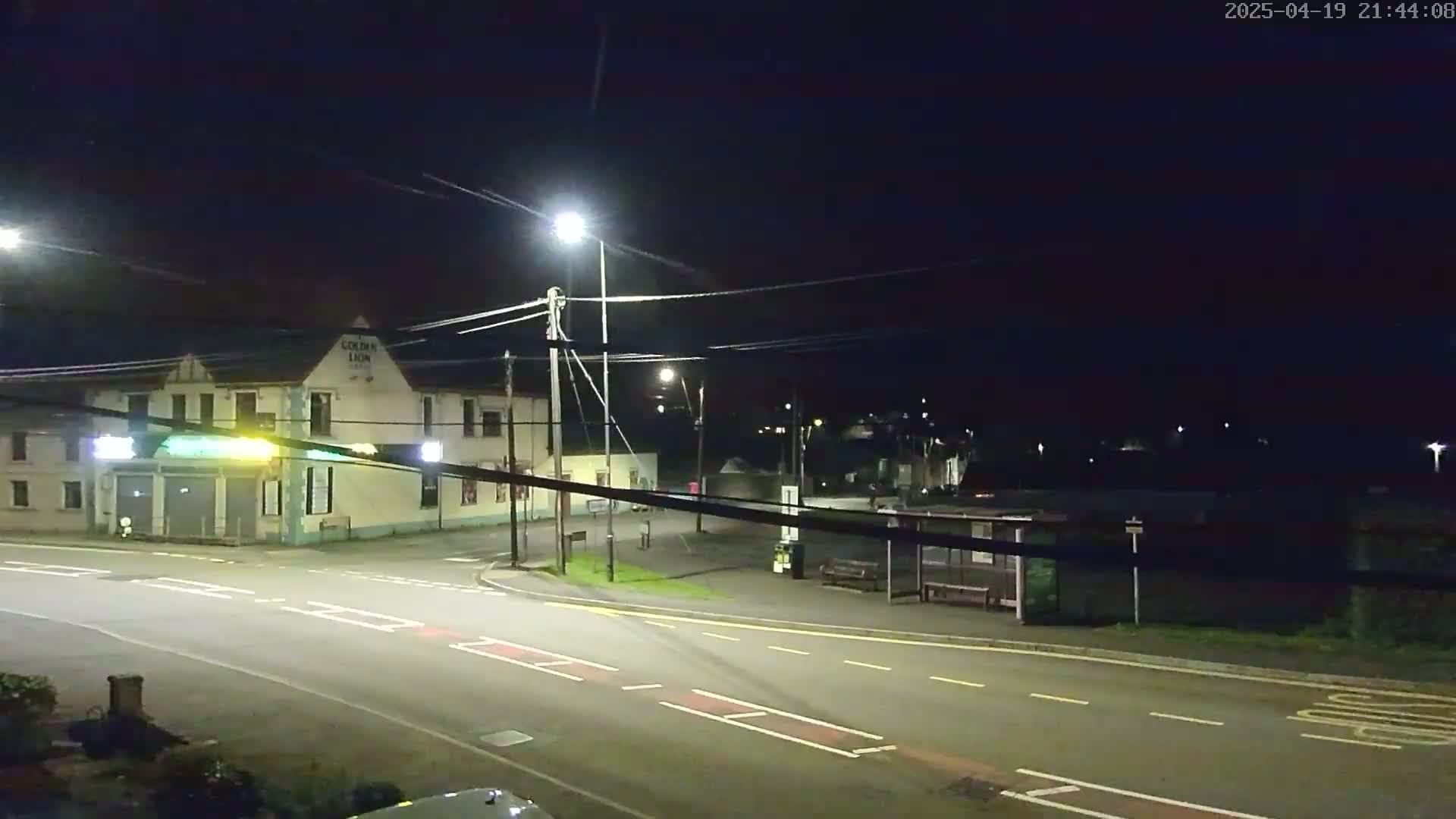 A nighttime street scene shows a building with illuminated signage, a bus stop, and a roadway under artificial light.