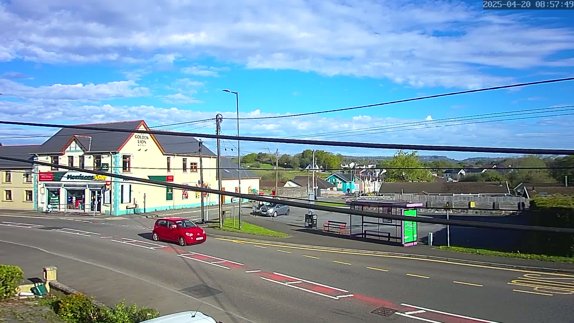 A partly cloudy blue sky overlooks a road intersection with a pub, a shop, a bus stop, and several cars, one of which is red.