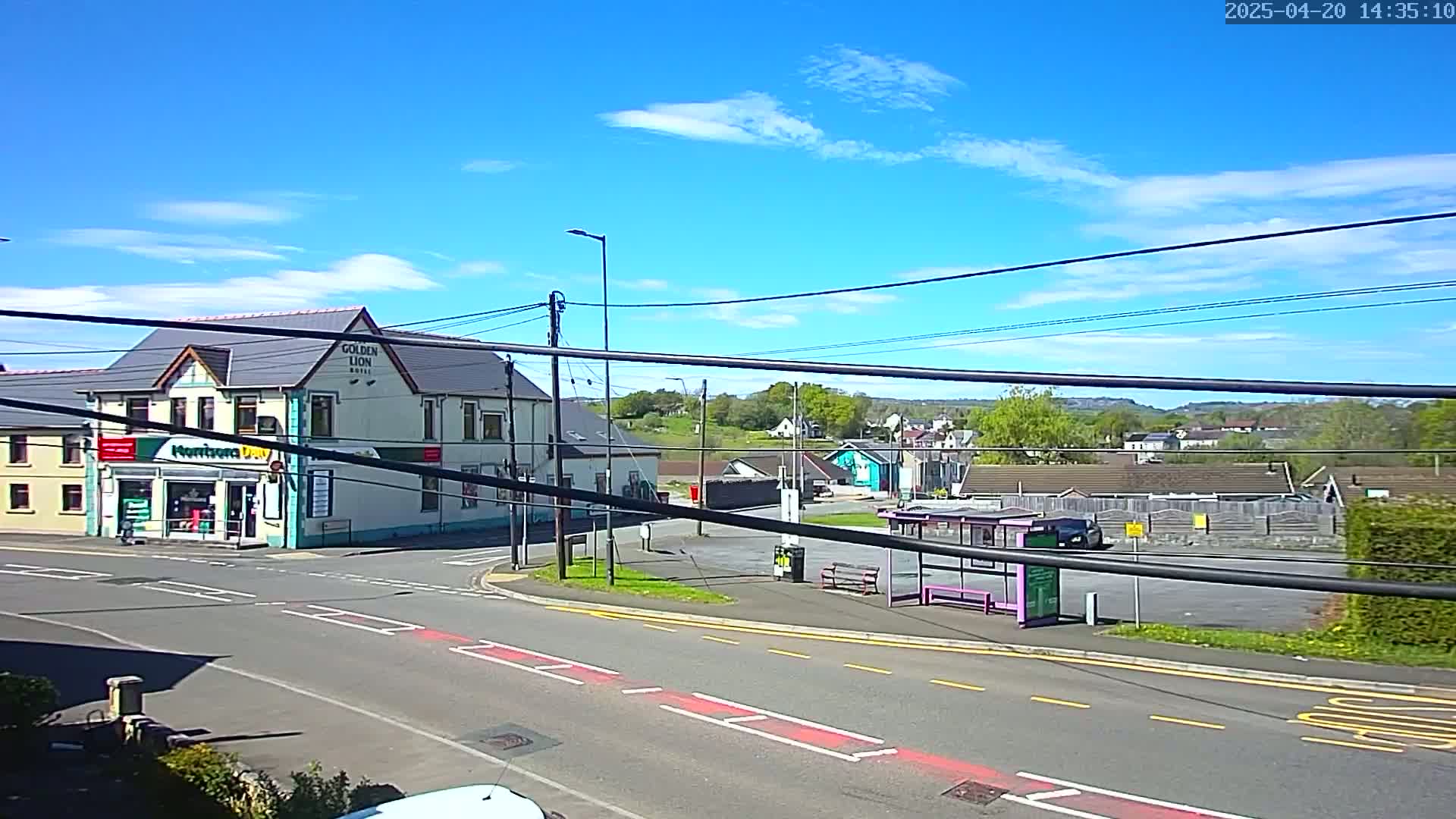 A sunny day reveals a street corner with a pub, a small shop, a bus stop, and residential buildings in the background.