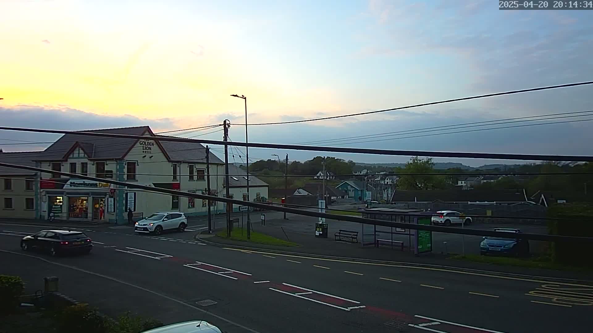 A street scene at dusk shows a two-story building, cars parked and driving on a road, and a bus stop, all under a partly cloudy sky.