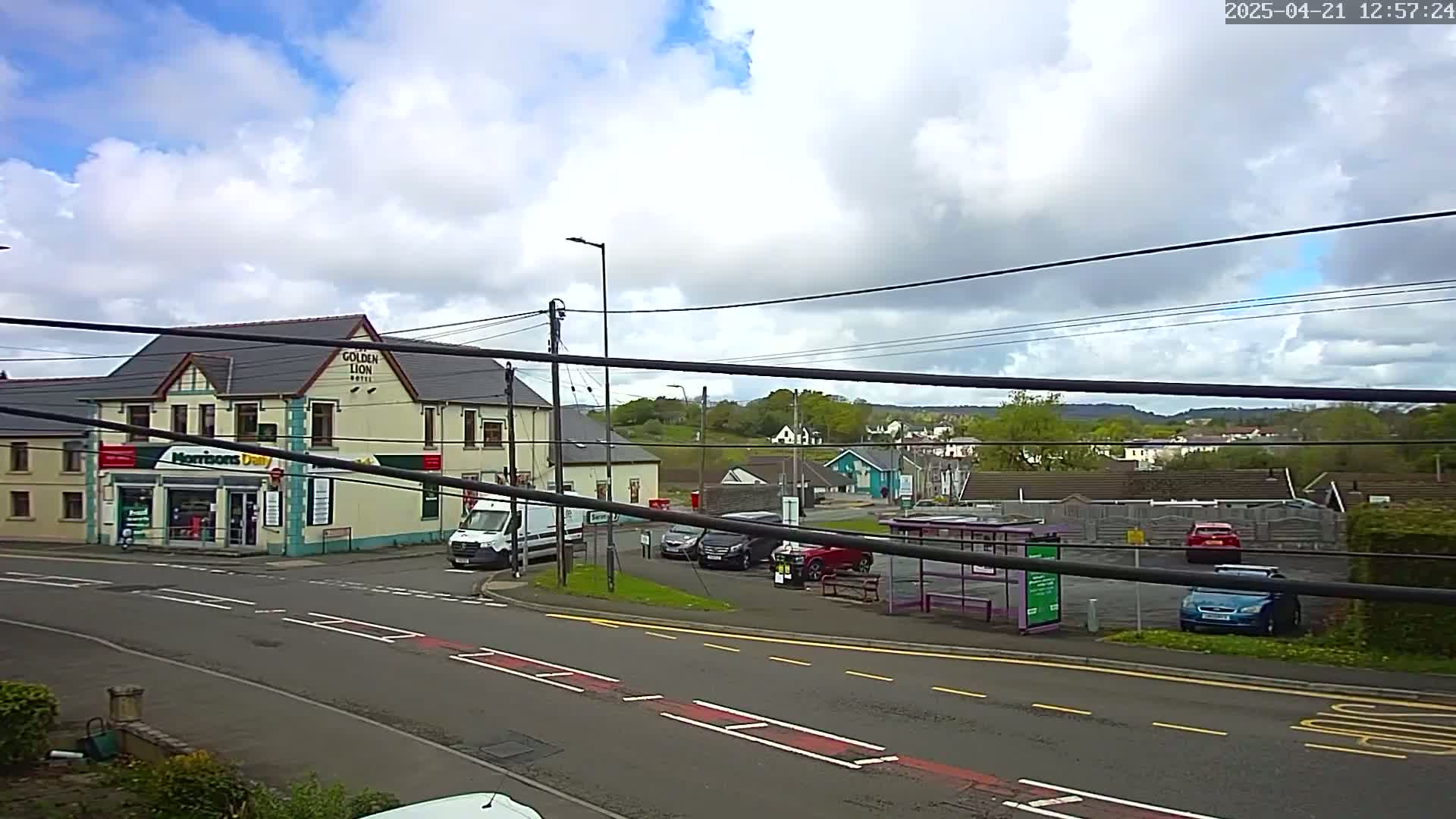 A partly cloudy day reveals a street scene with a hotel, a small store, several cars, and a bus stop, all situated near a crossroads.