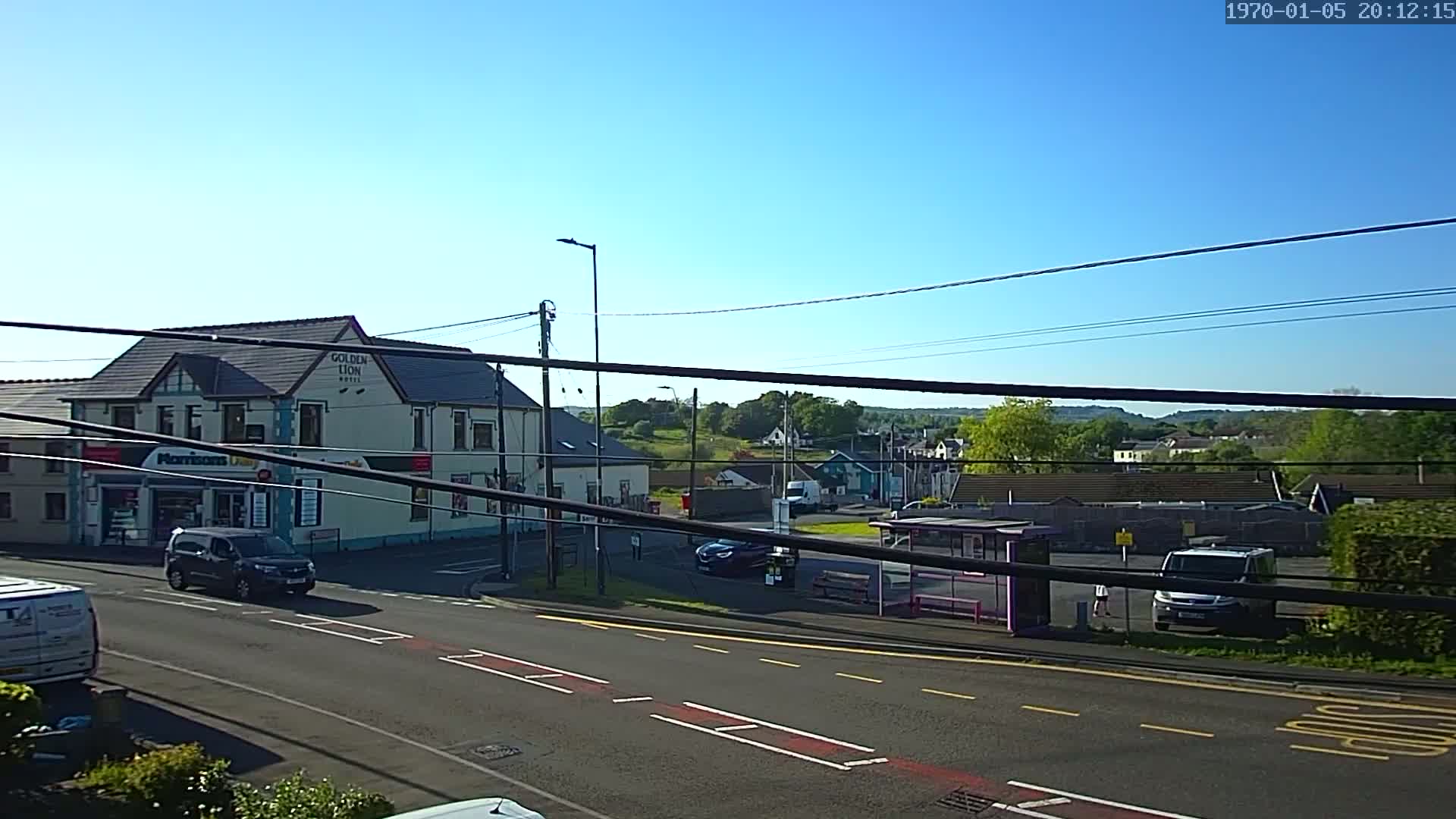A sunny day reveals a street scene with a two-story building, several vehicles, a bus stop, and a background of houses and greenery.