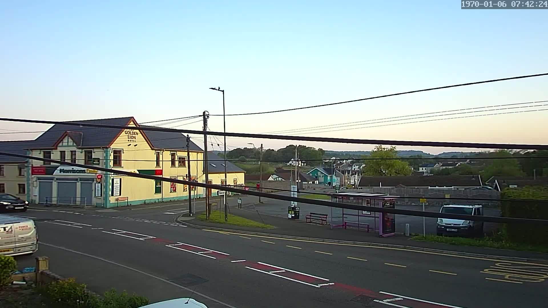 A yellow building stands at a road intersection, with a bus stop and other buildings visible in the background under a clear, twilight sky.
