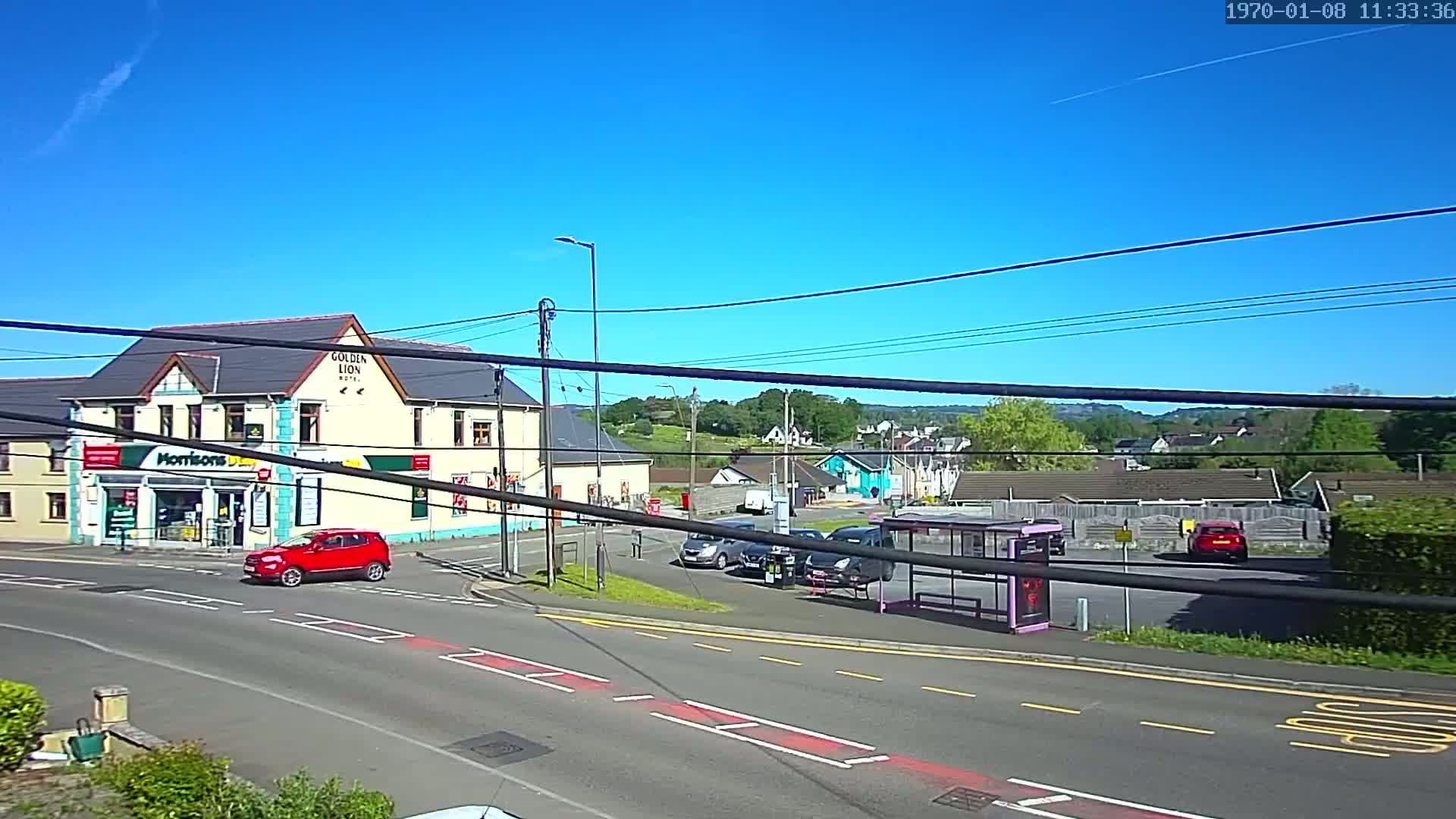 A sunny day reveals a street scene with a red car, a bus stop, several parked cars, and a building that includes a shop and hotel.