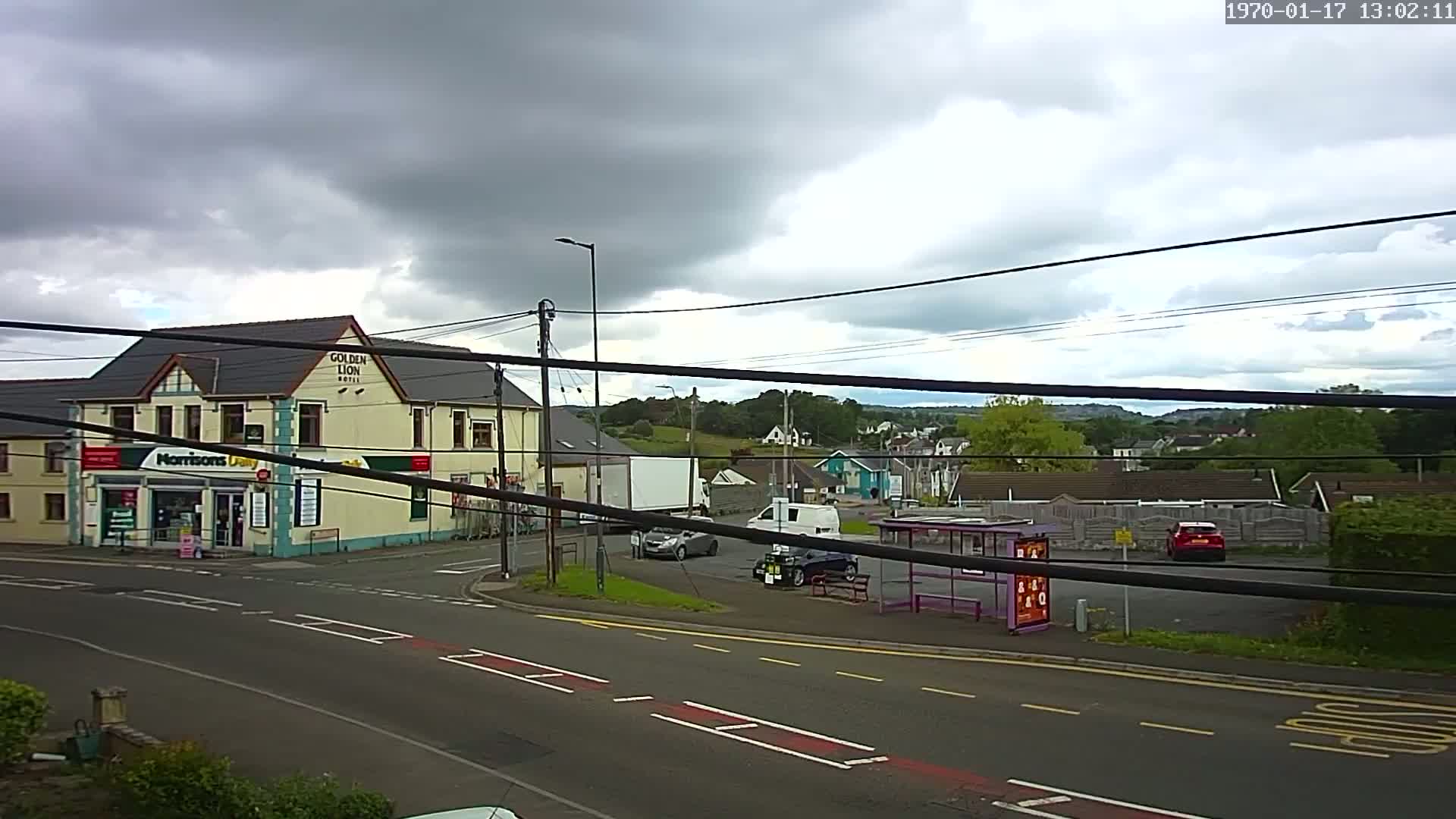 A cloudy day reveals a street scene with a hotel, a small shop, several cars, a bus stop, and a hilly landscape in the background.