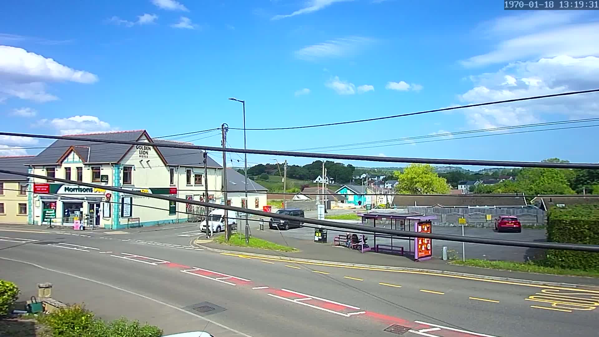 A sunny day reveals a street scene with a hotel, a shop, parked cars, a bus stop, and a small amount of green space in the background.