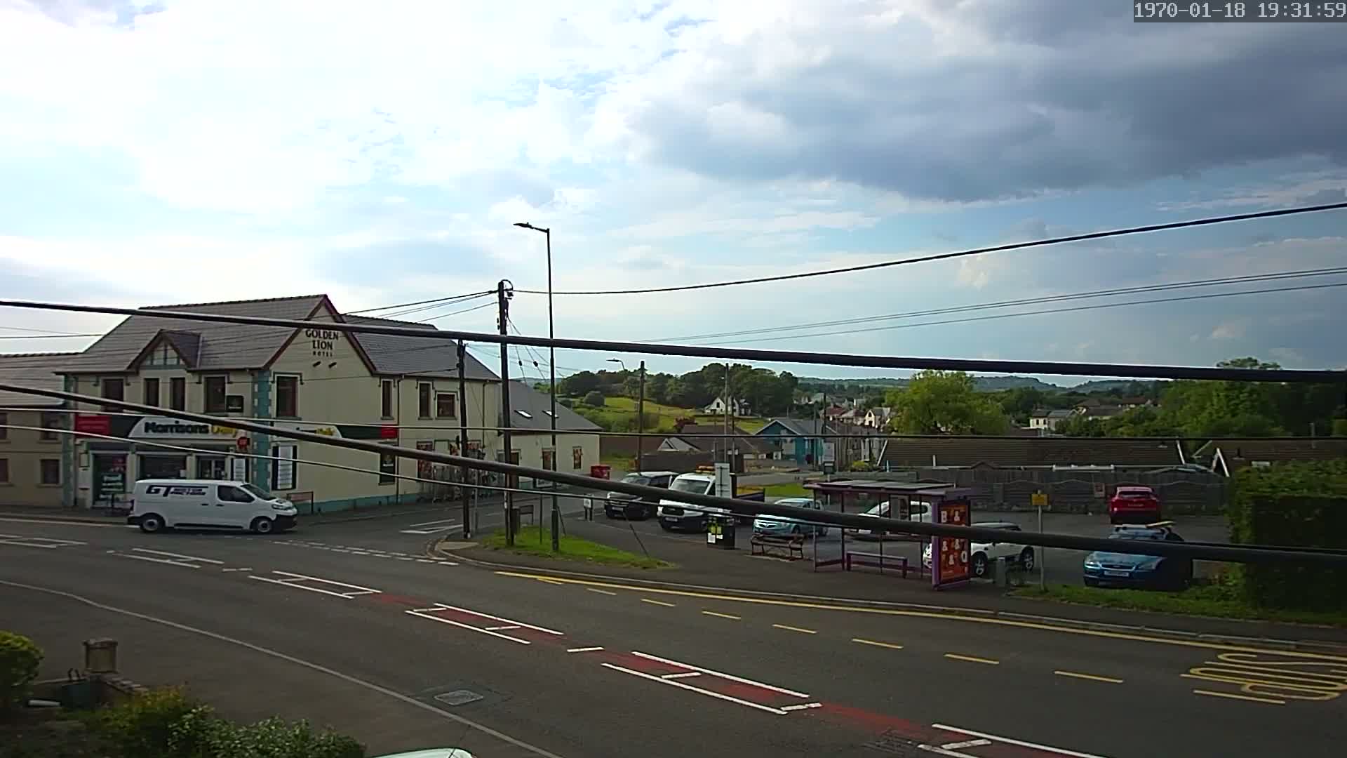 A partly cloudy day reveals a street scene with a hotel, a small shop, several vehicles, and a bus stop, all set against a backdrop of rolling green hills.