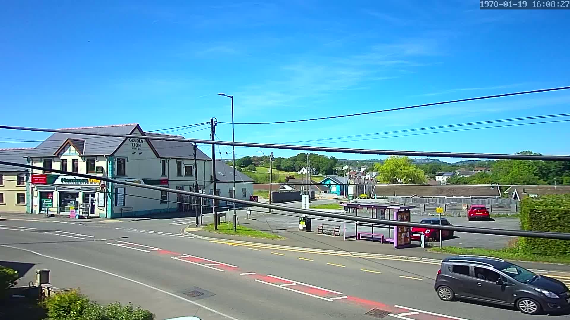 A sunny day shows a street corner with a two-story building, a bus stop, and several parked cars, all under a mostly clear blue sky.