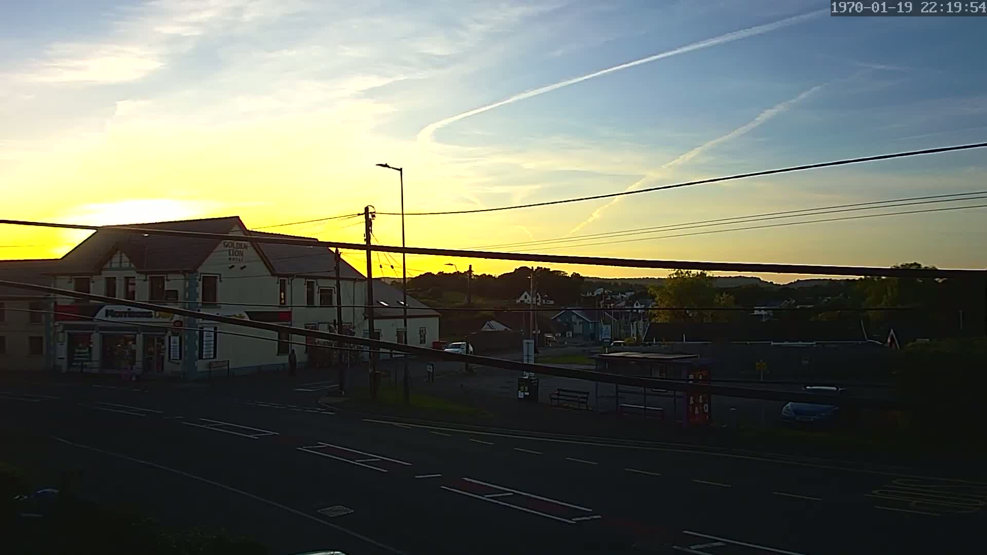 A sunset casts a yellow glow over a street scene with buildings, power lines, and a few cars, under a mostly clear sky with contrails.