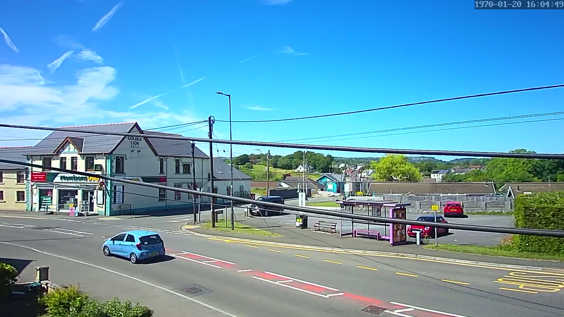 A blue car drives past a corner intersection with a hotel and a small shop under a clear blue sky with contrails.