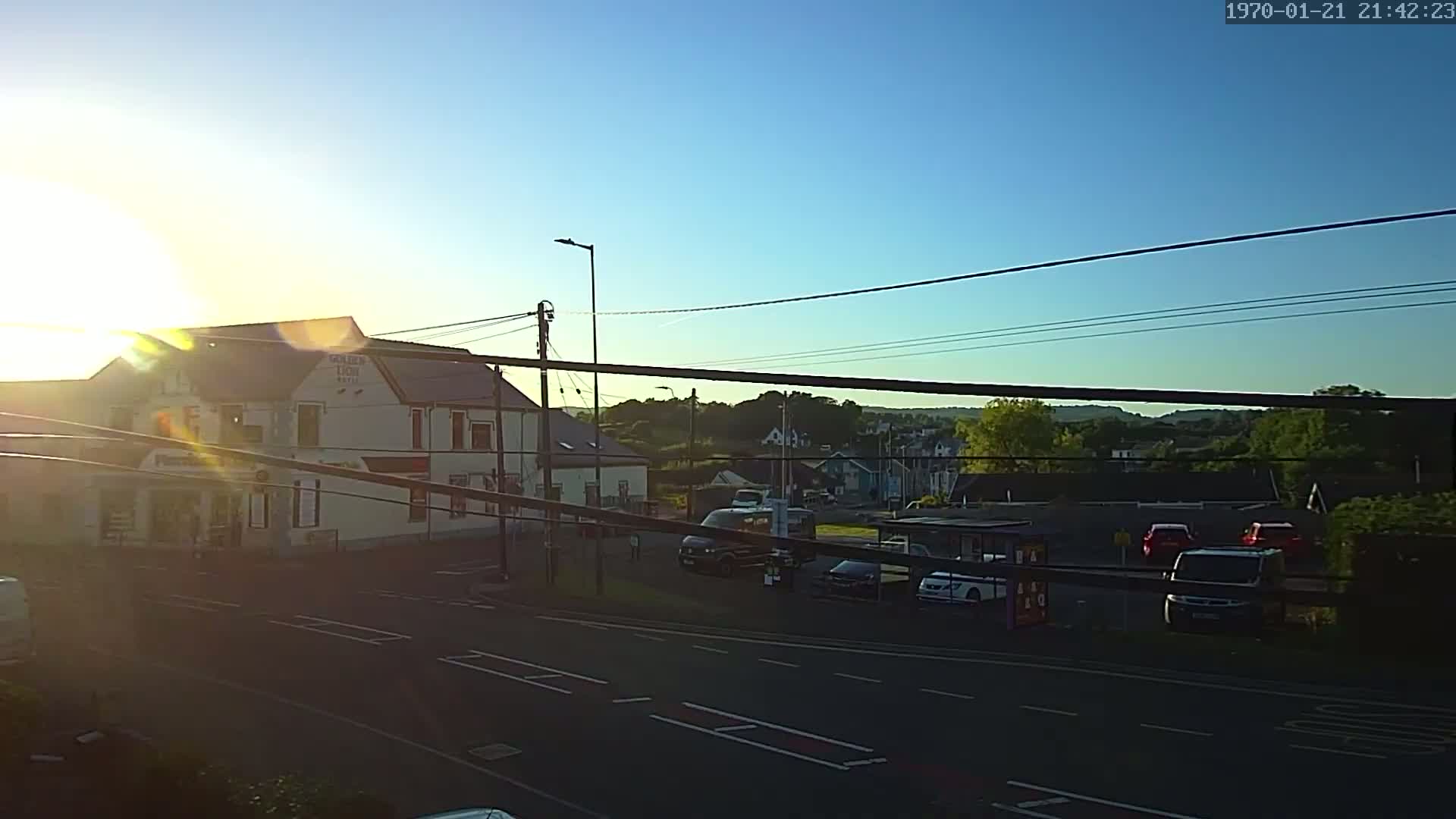A brightly lit sunrise shines over a street scene with buildings, parked cars, and a few vehicles on the road, all under a clear sky.