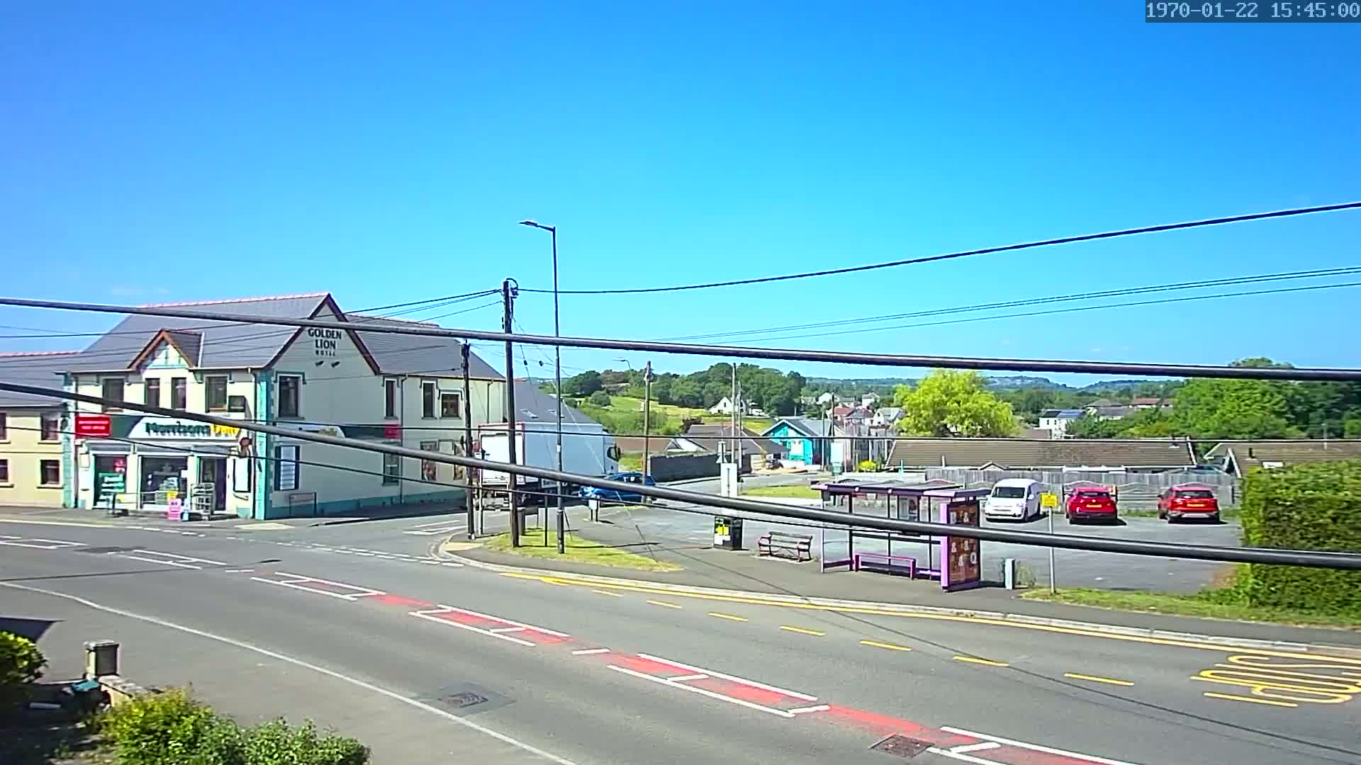 A sunny day reveals a street scene with a two-story building housing a store and a hotel, a bus stop with several parked cars, and a background of greenery and other buildings under a clear blue sky.
