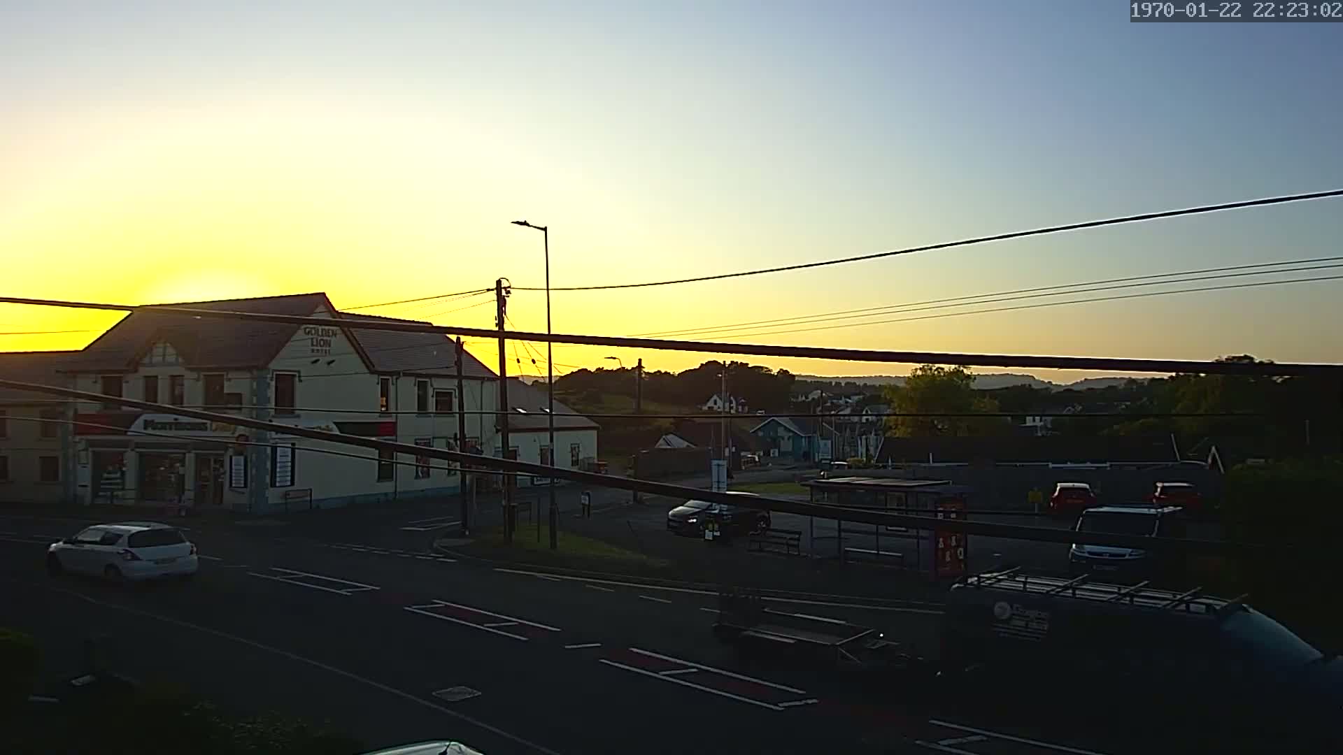 A sunset illuminates a street scene with buildings, cars, and power lines, showing a mostly clear sky.