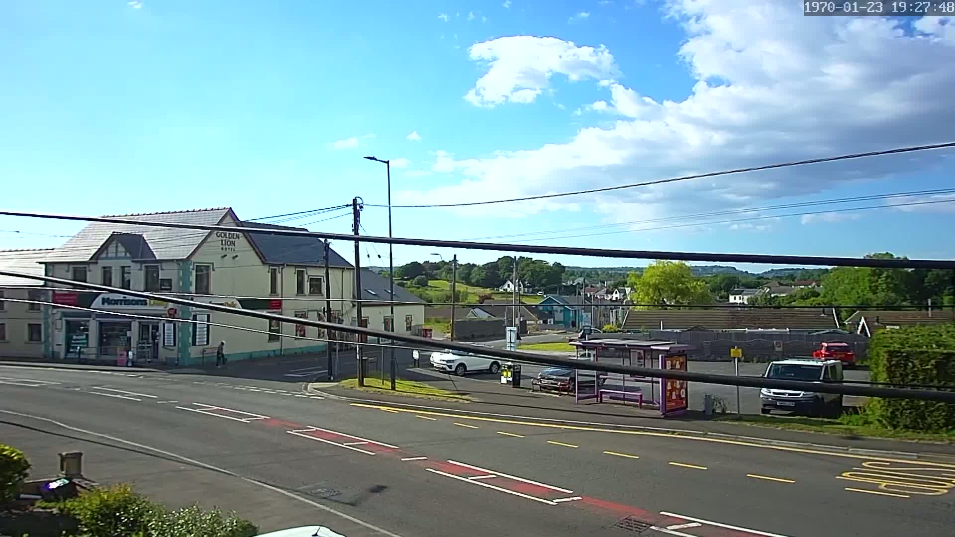 A partly sunny day reveals a street scene with a hotel, a small shop, several cars, a bus stop, and a hilly background.