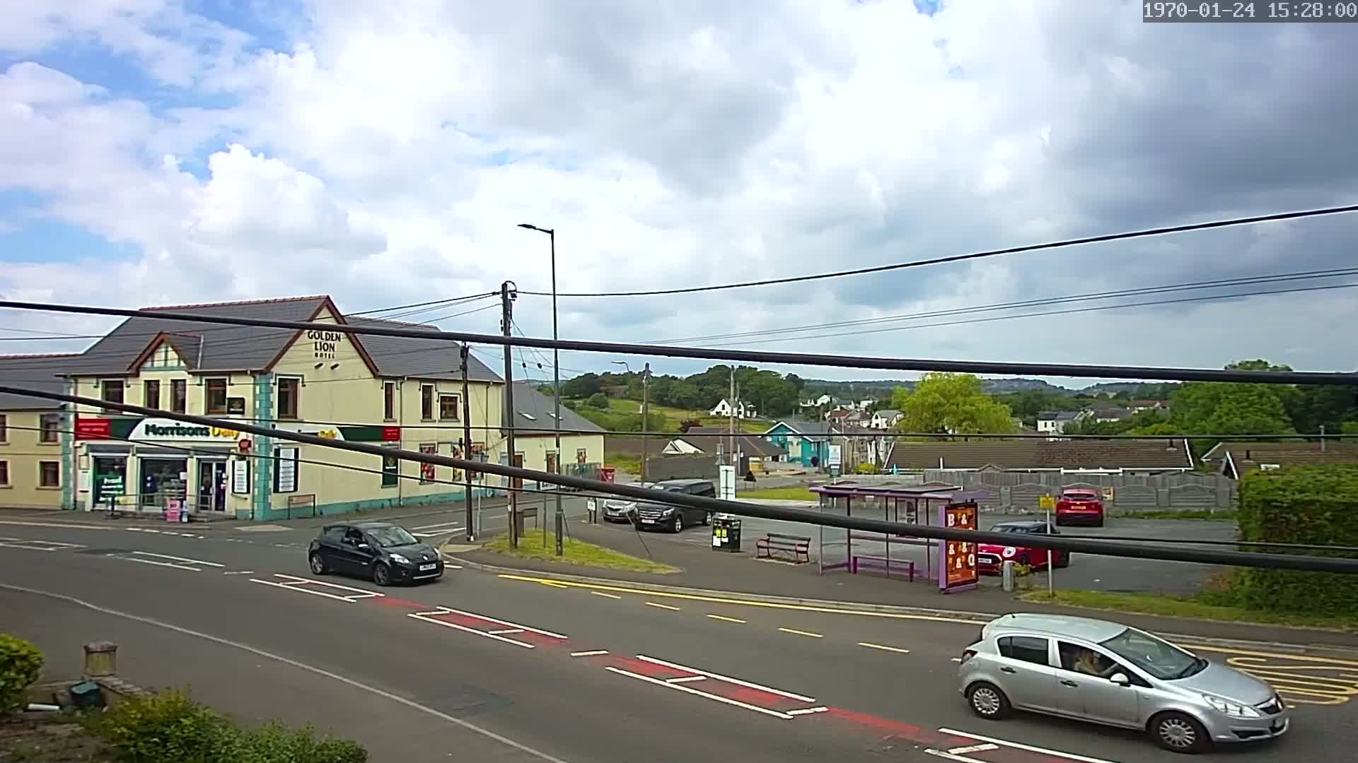 A partly cloudy day shows a street scene with cars, a bus shelter, a small supermarket, and a hotel in the background, all under a network of power lines.