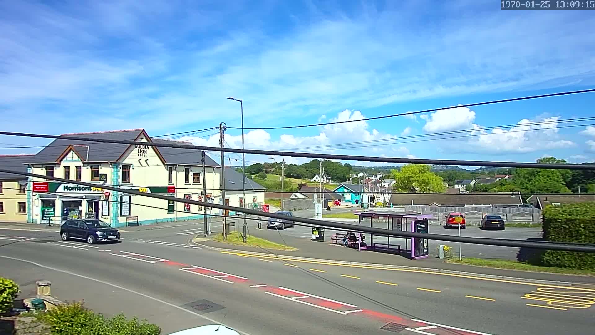 A sunny day reveals a street scene with a pub, a small store, several cars, a bus stop with a person sitting inside, and residential buildings in the background.