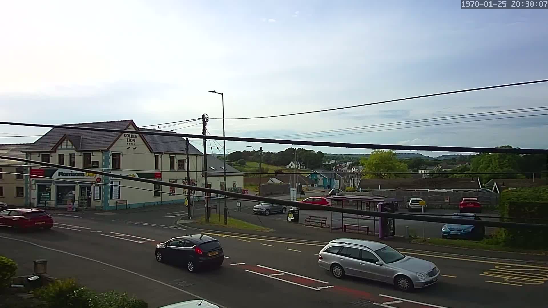 A partly cloudy day reveals a street scene with several cars and a bus stop near a hotel and small store, all surrounded by low-lying buildings and green hills in the background.