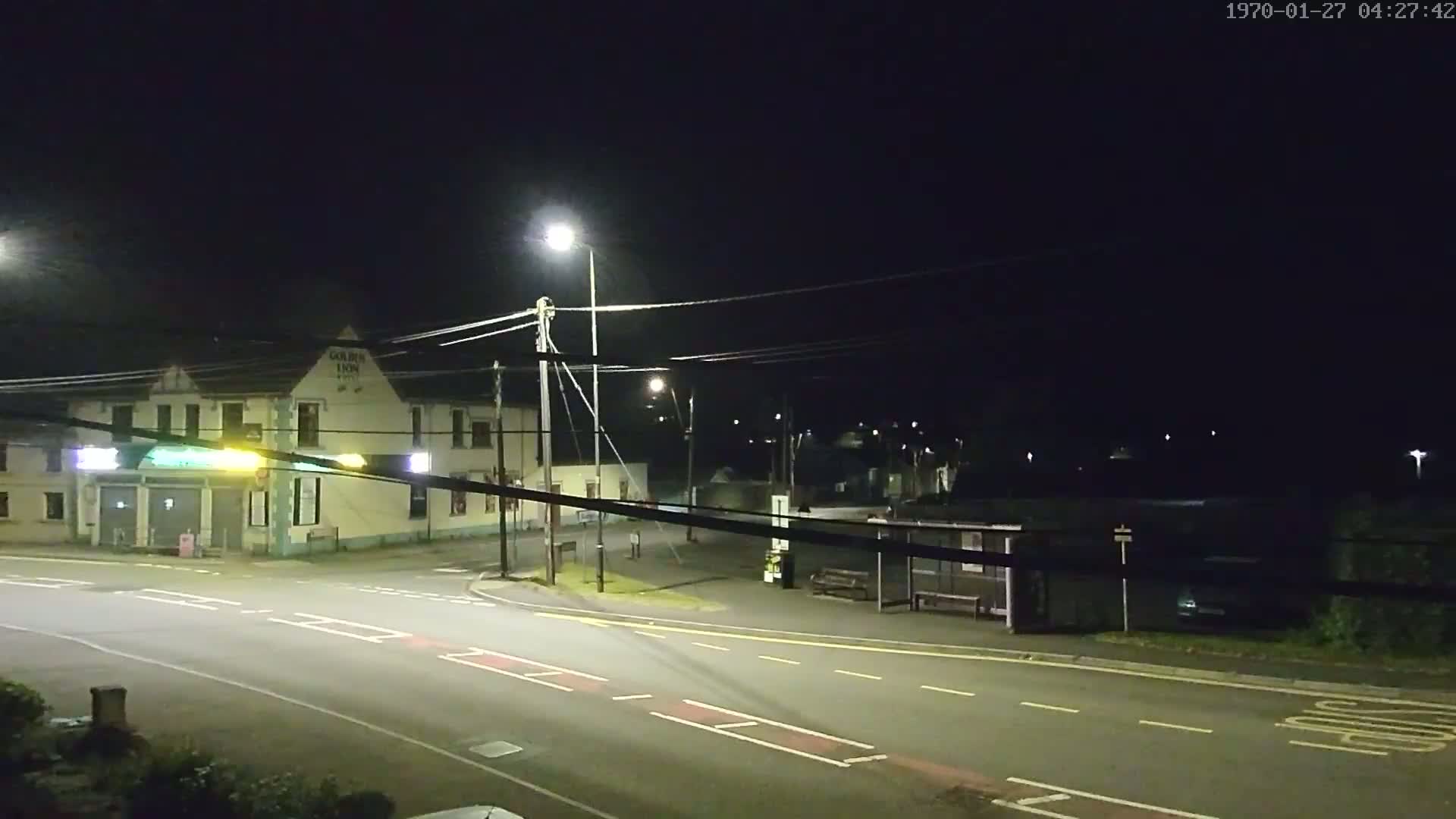 A nighttime street scene shows a building with lights on, streetlights illuminating a road intersection with a bus stop, and a dark sky.