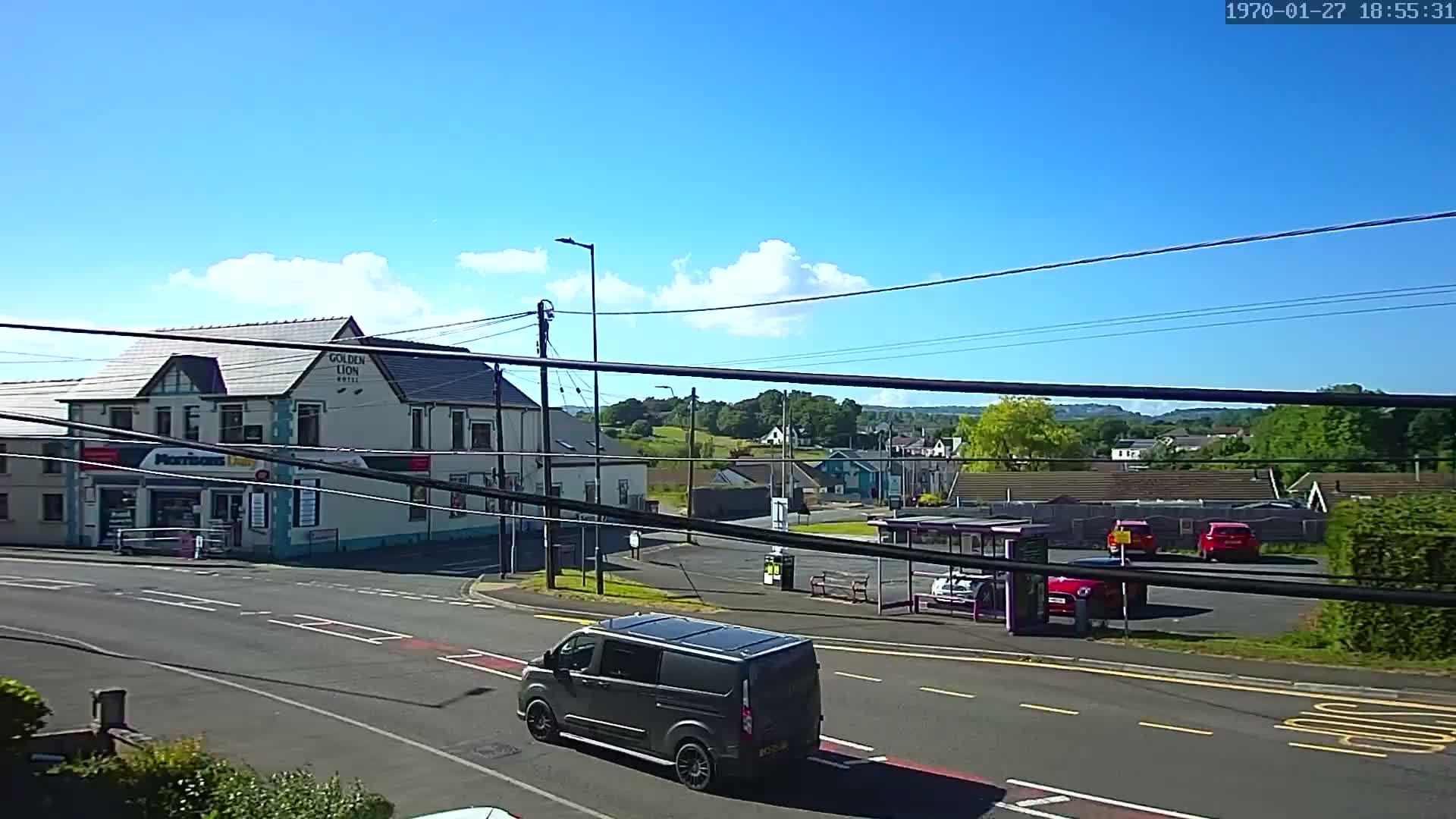 A dark-grey van drives past a crossroads on a sunny day with some scattered clouds, near a hotel and a bus stop.
