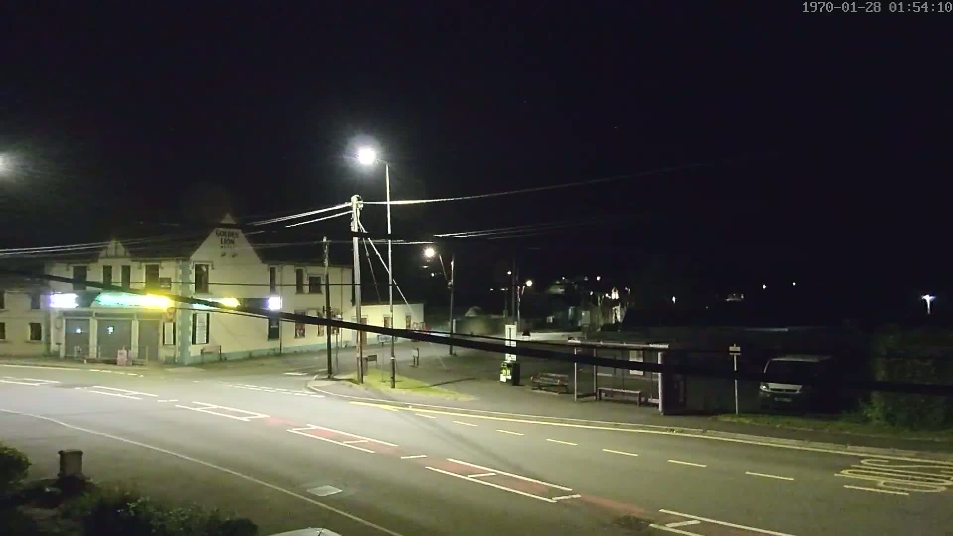 A nighttime street scene shows a road intersection with a building, a bus shelter, and a van under artificial light.