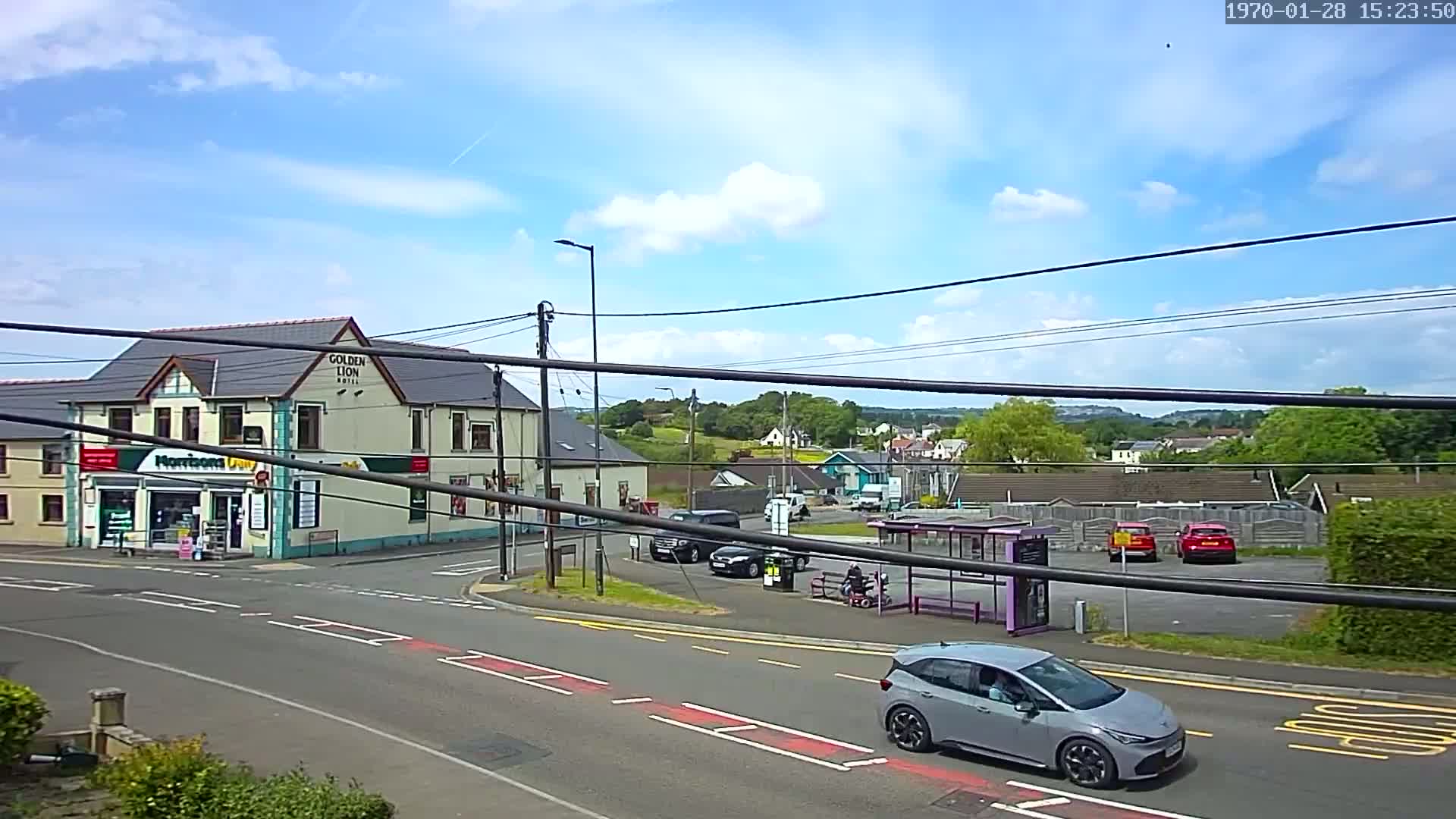 A partly sunny day shows a street scene with a hotel and small shop, cars parked and driving, and a bus stop with a person sitting.