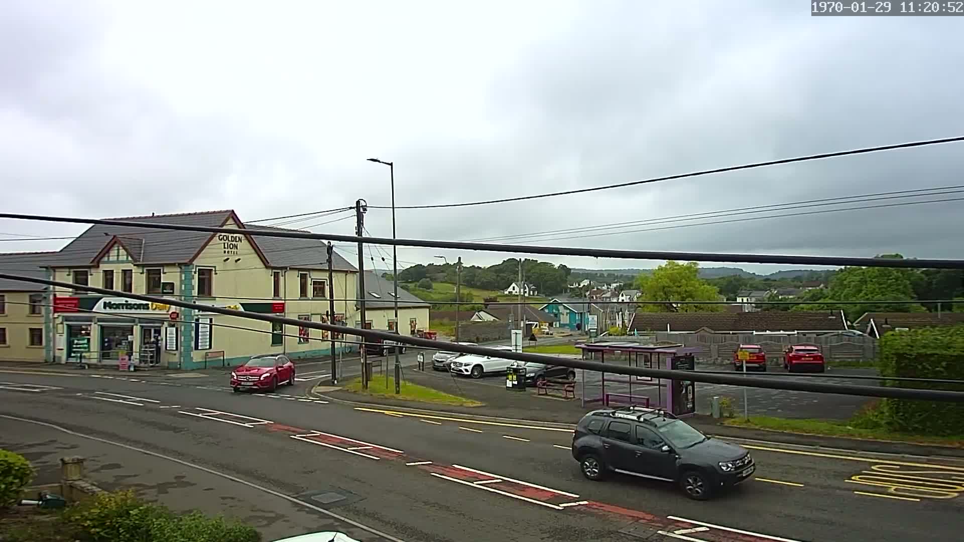 A street scene on an overcast day shows a crossroads with several cars, a bus stop, a hotel, and a small shop.