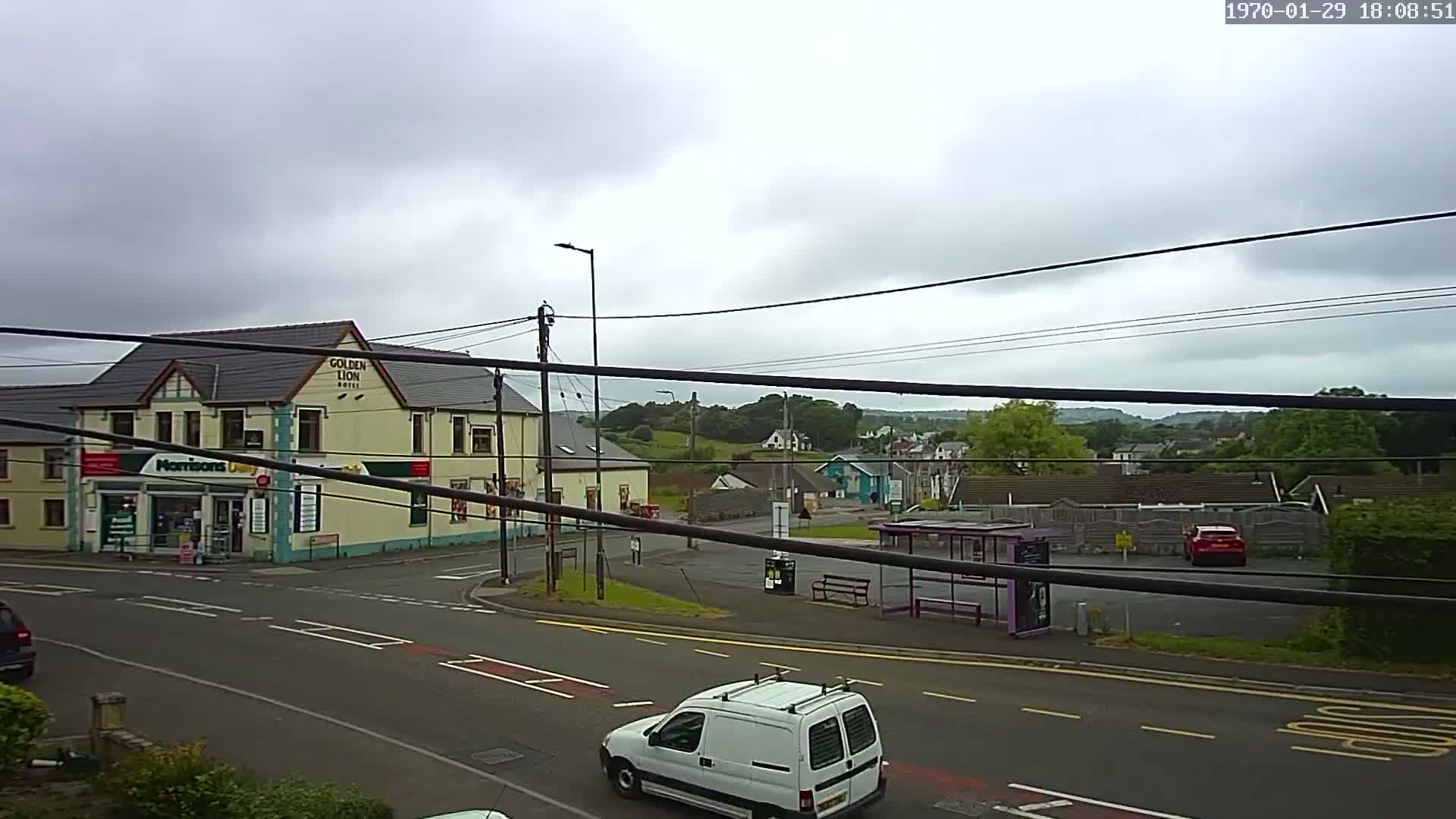 A cloudy day reveals a street scene with a hotel, a small shop, a bus stop, several cars, and a van, all set against a backdrop of low-lying hills and houses.