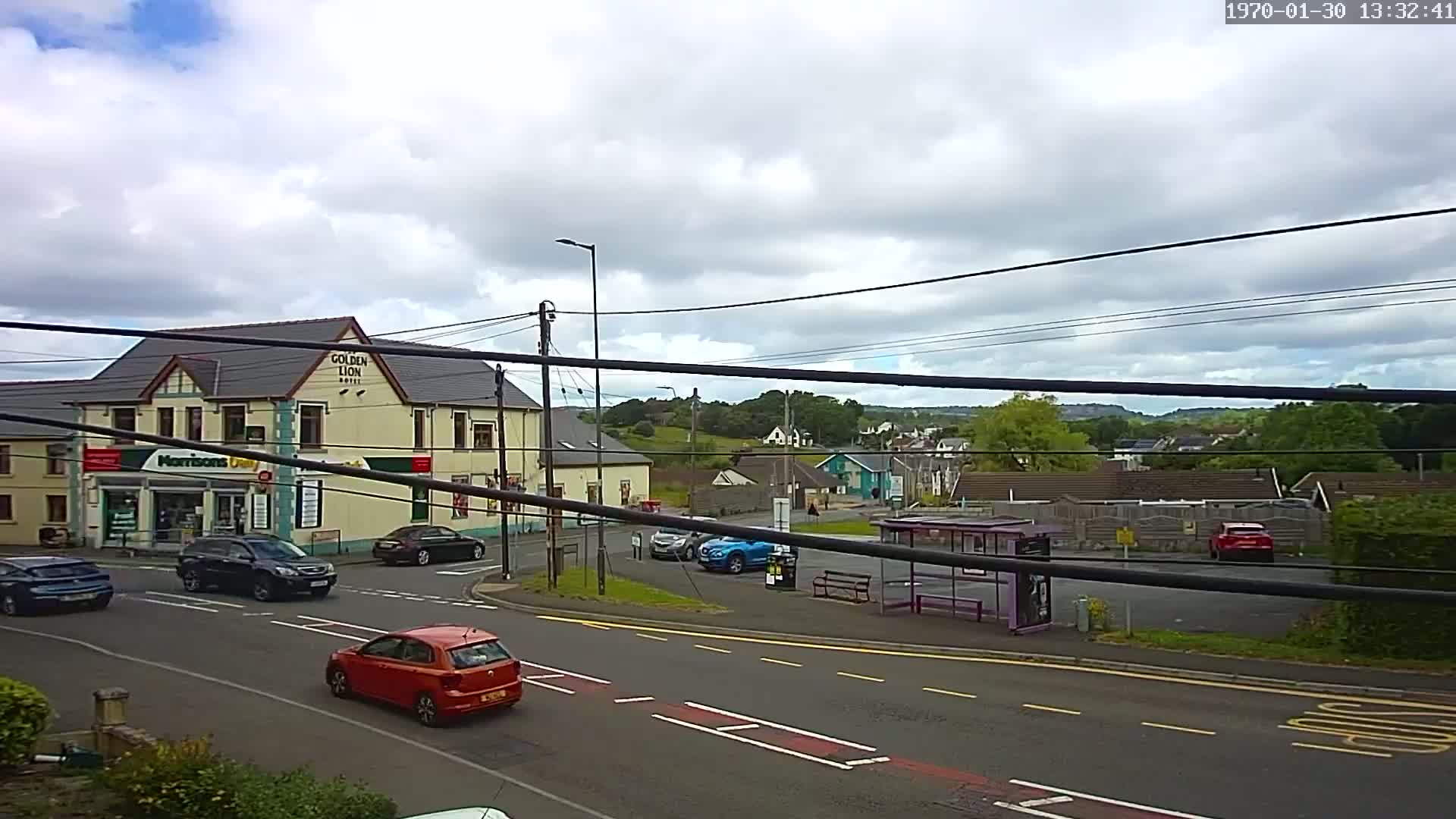 A partly cloudy day reveals a street scene with several cars, a bus stop, a hotel, and a small shop, all situated near a hilly landscape.