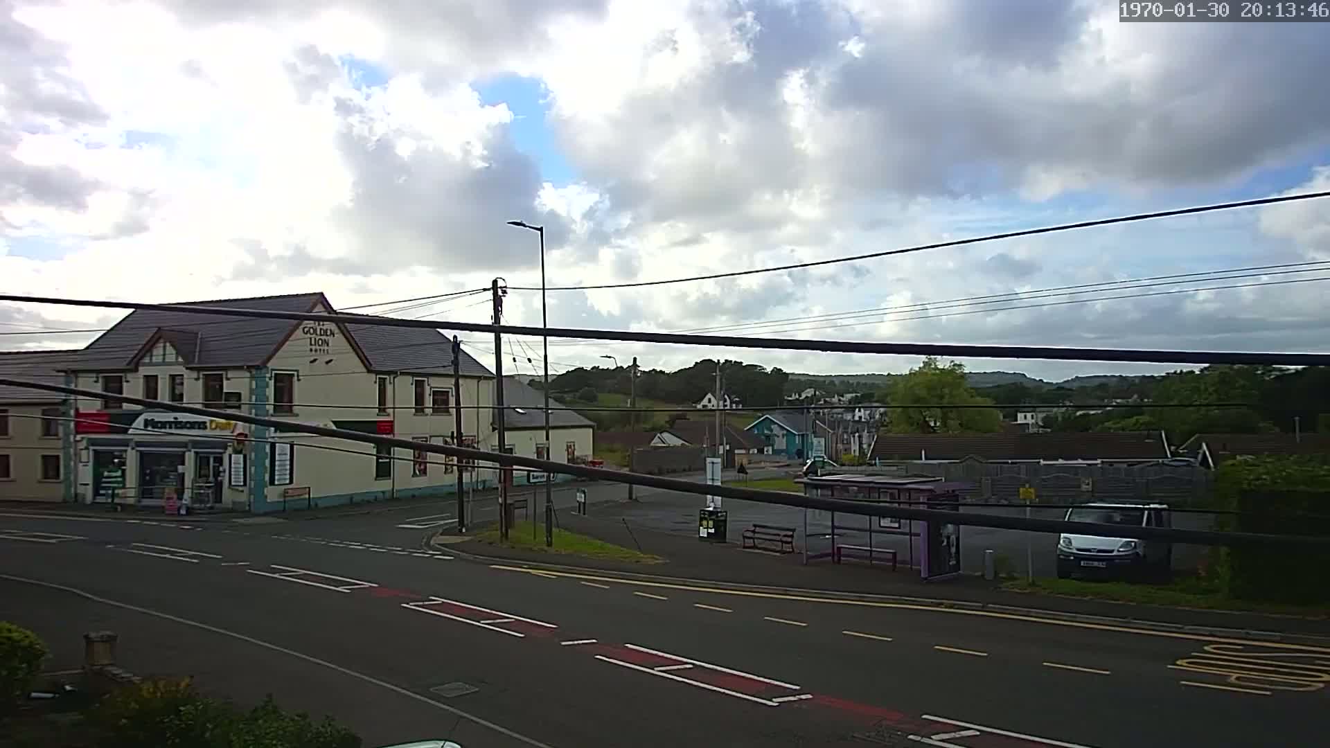 A partly cloudy day shows a street scene with a hotel, a small shop, a bus stop, and a few cars, all surrounded by residential buildings and trees.