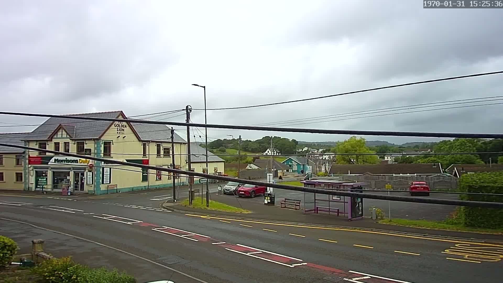 A street scene on an overcast day shows a two-story building with a shop on the ground floor, a bus stop, several cars in a parking lot, and trees and houses in the background.