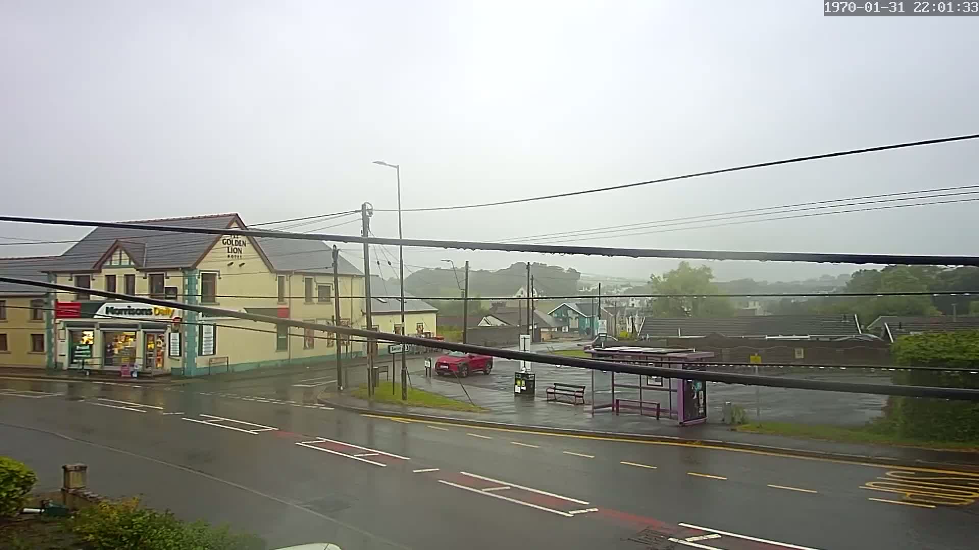 A wet, overcast street scene shows a two-story building with a small shop next to it, a bus stop, and a red car parked in a small lot.