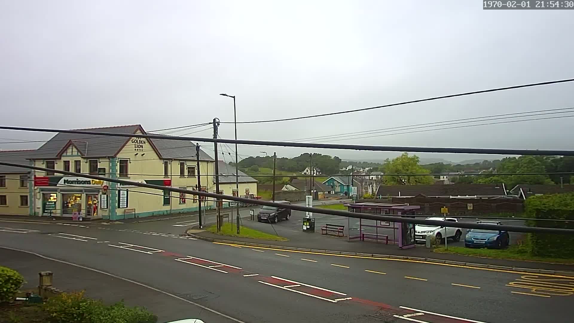 A wet, overcast day reveals a street scene with a hotel and shops, a bus stop with cars parked nearby, and a residential area in the background.