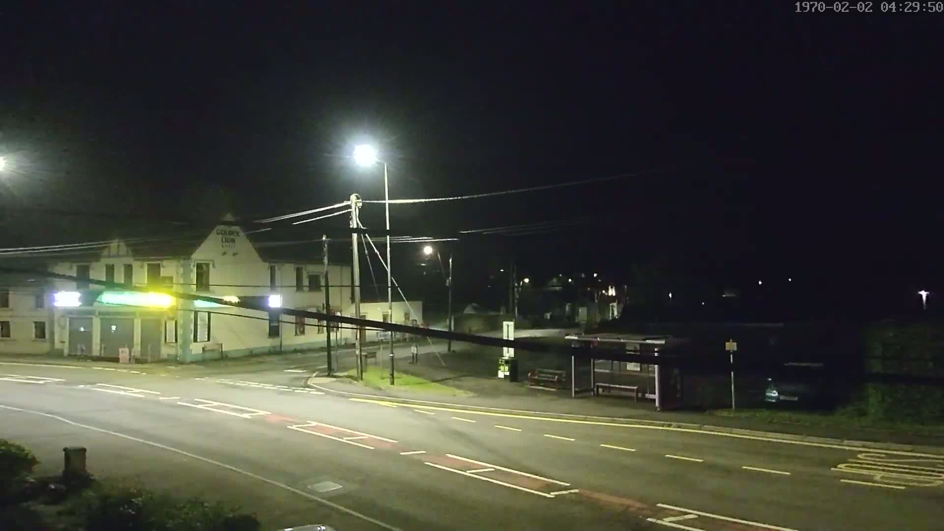 A nighttime street scene shows a road intersection with a building, a bus shelter, and streetlights illuminating the area under a dark sky.